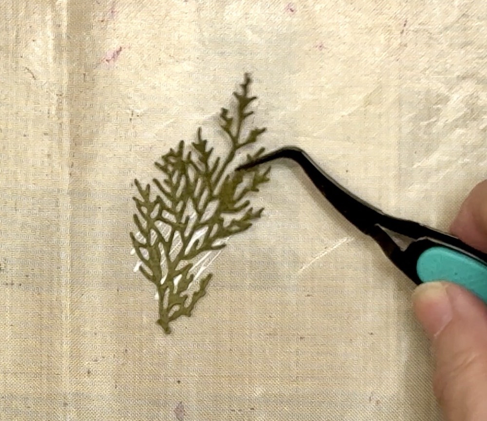 Screen shot showing dabbing the greenery branch into the glue on the Teflon mat to aid in getting just enough glue to adhere the branch to the card front. 