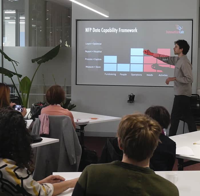 a person presenting a table on a screen in an office. People are sitting at white tables listeninng.