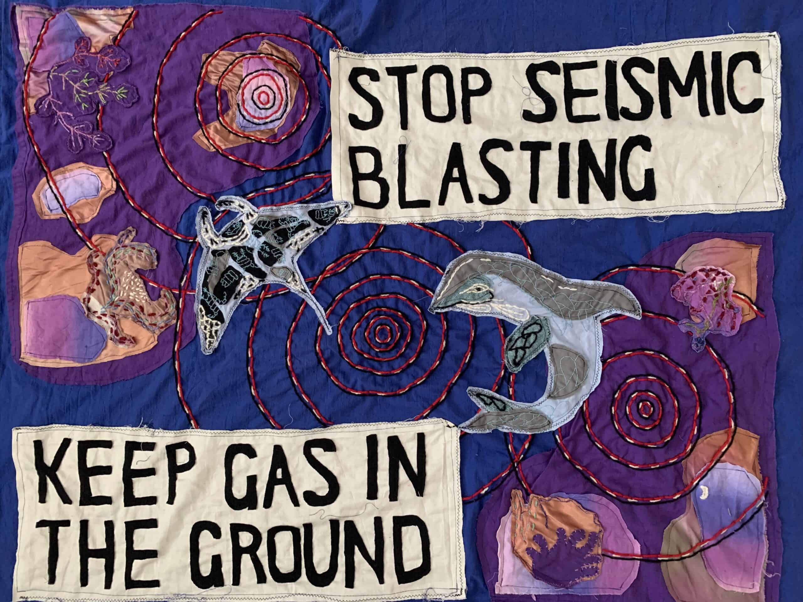 ID: A hand sewn banner with corals, a manta ray and a dolphin with black and red lines radiating out diagonally across the banner, criss-crossing over the wildlife. The text is arranged in the top right and bottom left corners and reads “STOP SEISMIC BLASTING KEEP GAS IN THE GROUND”