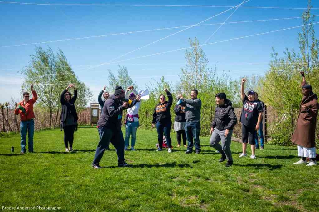 Photo of participants in an outdoor activity. They hold a web of string bove their heads. The scene is a grassy backyard with blue sky behind.