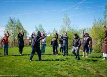 Photo of participants in an outdoor activity. They hold a web of string bove their heads. The scene is a grassy backyard with blue sky behind.