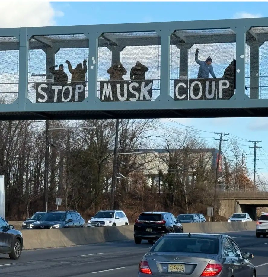 People standing on a covered bridge above a busy road. There is a sign for the the car drivers to read. The sign reads 'Stop Musk Coup'.