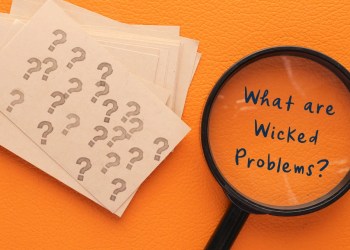 A magnifying glass next to a pile of cards with stamped question marks. Inside the magnifying glass is the text 'What are wicked problems?'.