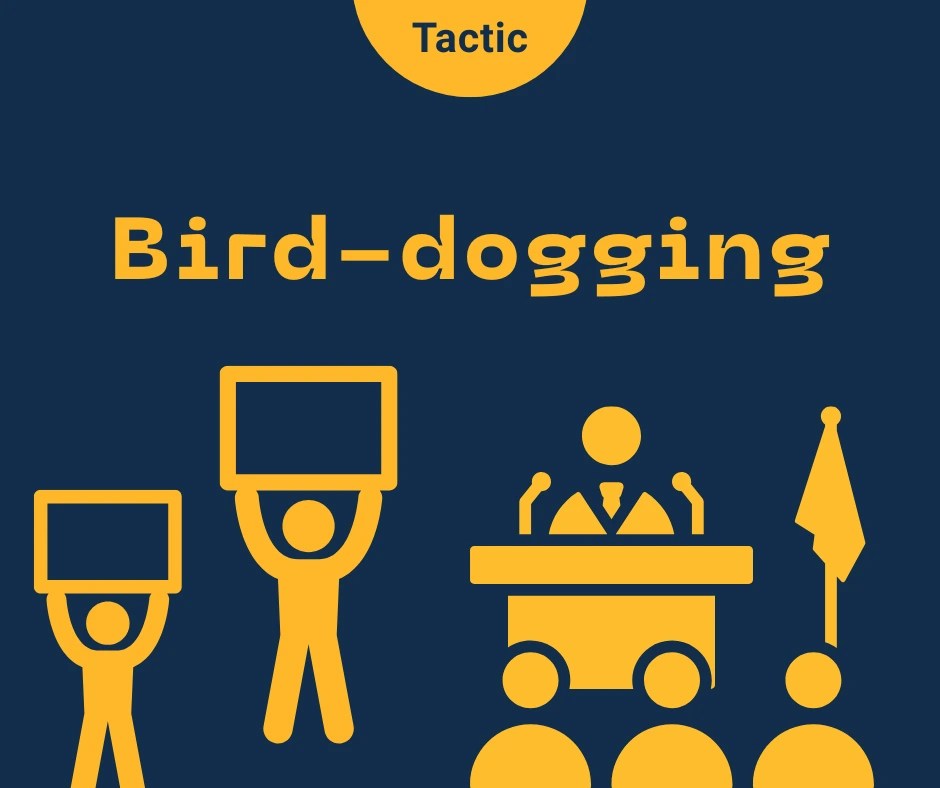 Title reads 'Bird-dogging'. A person speaks at a lecturn to an audience. Two people are at the side holding placards up above the heads.