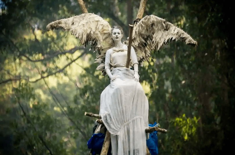 A woman in an angel costume sits atop a tripod in Tasmania's old growth forest.