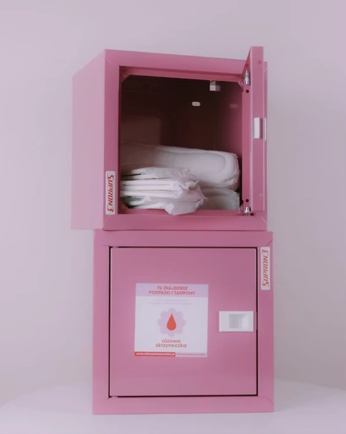 2 metal square pink locker boxes stacked on top of each other. The top one has its door open and is filled with white menstrual pads.