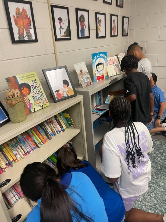 group of upper elementary students gathered around a neat and inviting bookshelf looking at books.