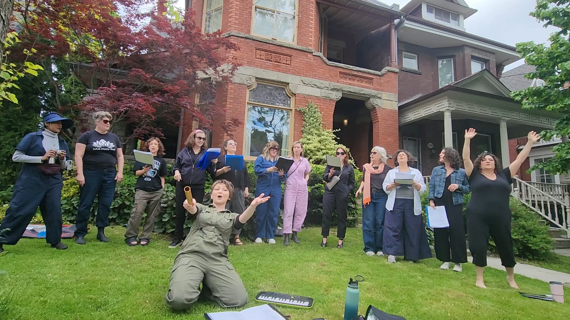 An image of 13 women on the front lawn of a brick house. They are singing from song sheets. Two of the women are gesturing with wide open arms.