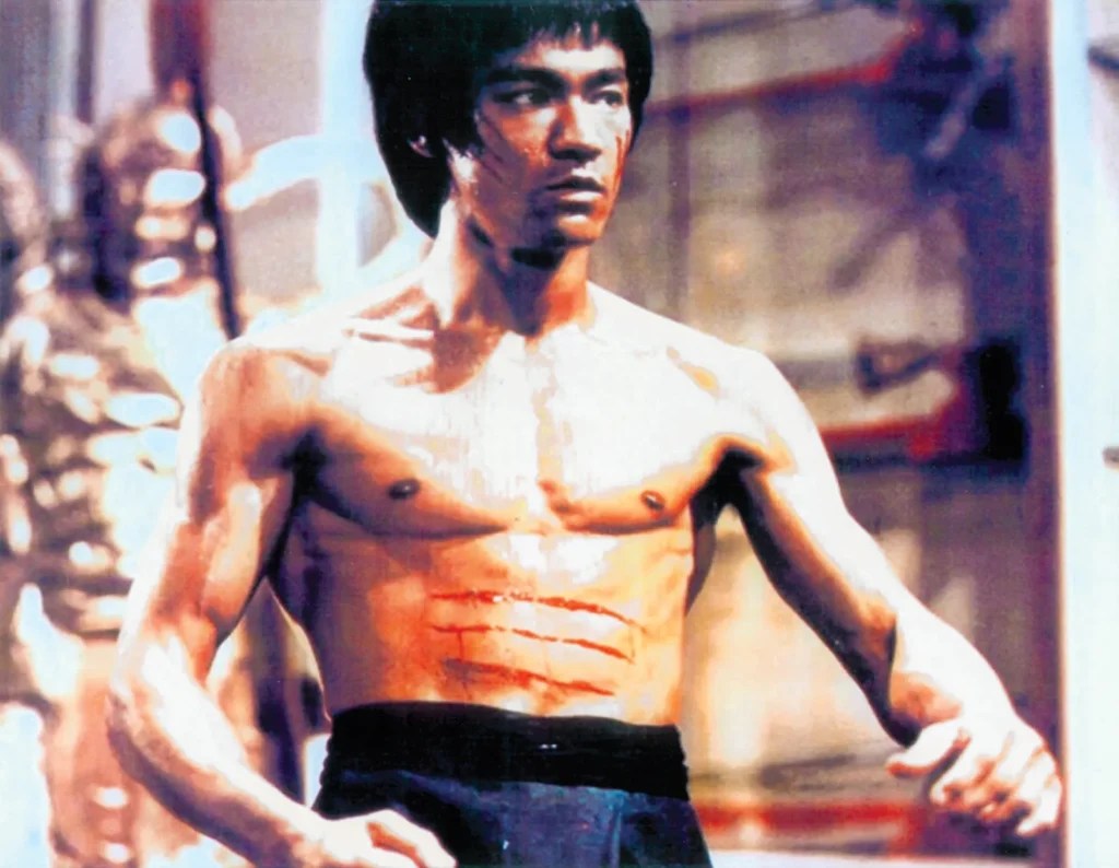 Un hombre sin camiseta con marcas de arañazos en el pecho adopta una postura de artes marciales en el interior, con un aspecto concentrado que recuerda a los luchadores legendarios y despierta la curiosidad por CÓMO MURIÓ BRUCE LEE.