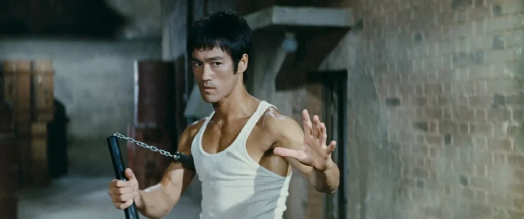Un hombre con camiseta blanca de tirantes sostiene un nunchaku en una mano y levanta la otra en una postura de artes marciales contra una pared de ladrillo, evocando momentos clásicos de CÓMO MURIÓ BRUCE LEE.