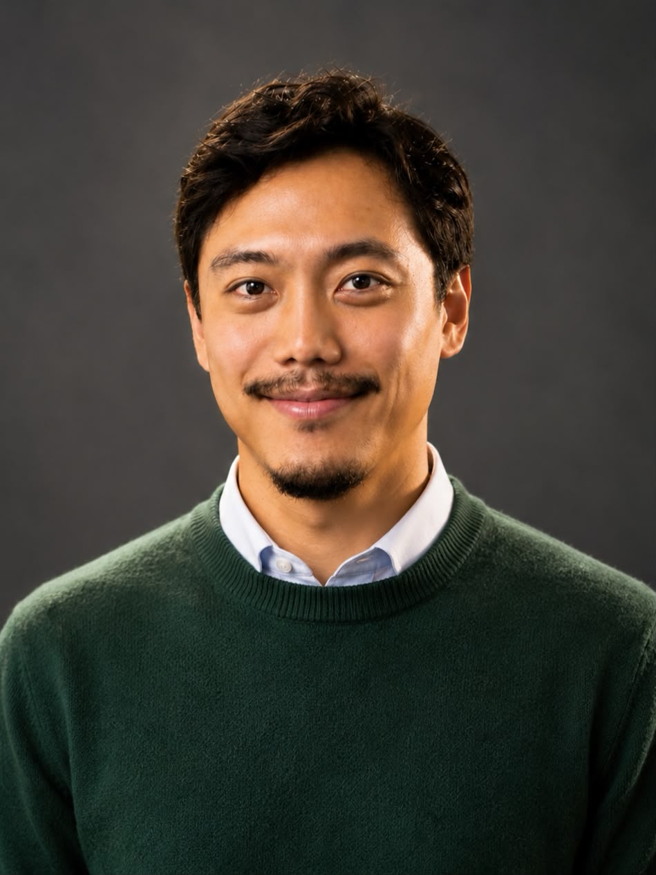 A portrait of a smiling man with short black hair, wearing a green sweater and a light blue shirt, against a dark background.