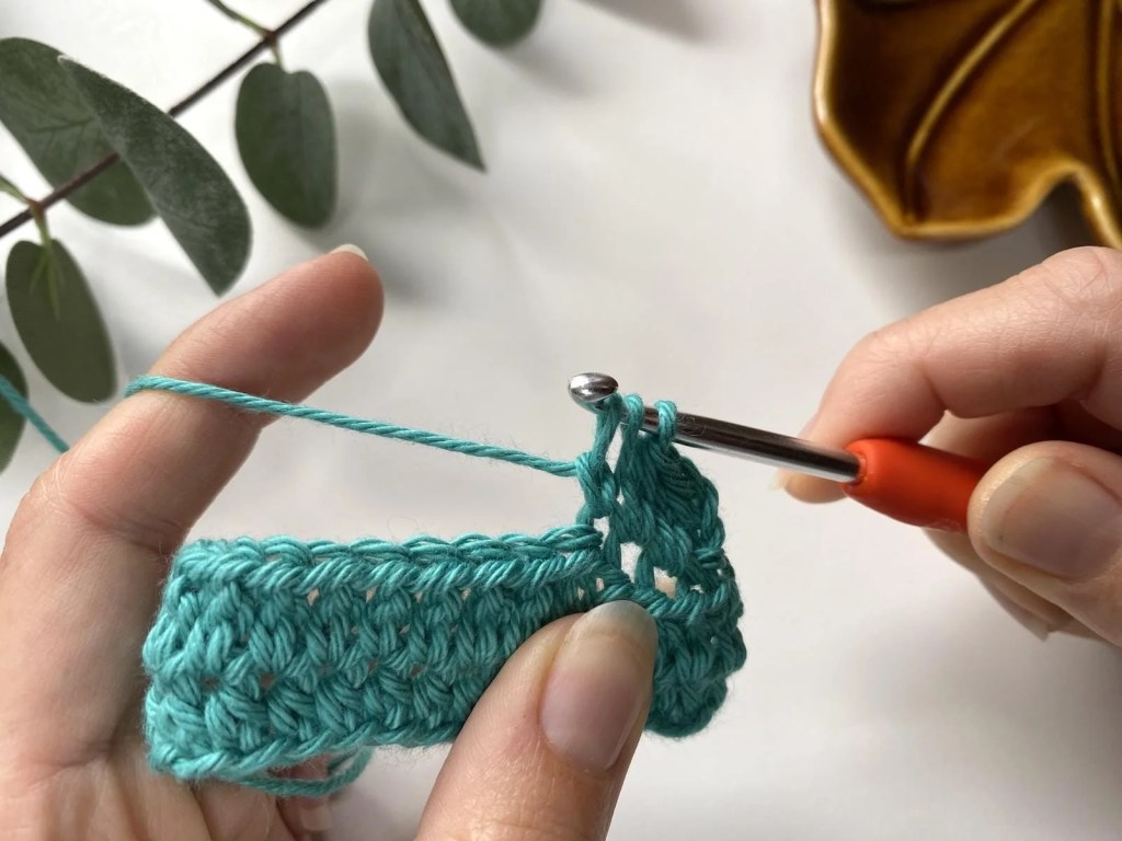 yarn over and pull through 1 loop is being demonstrated by 2 hands with a crochet hook and teal yarn making a beginner crochet stitch