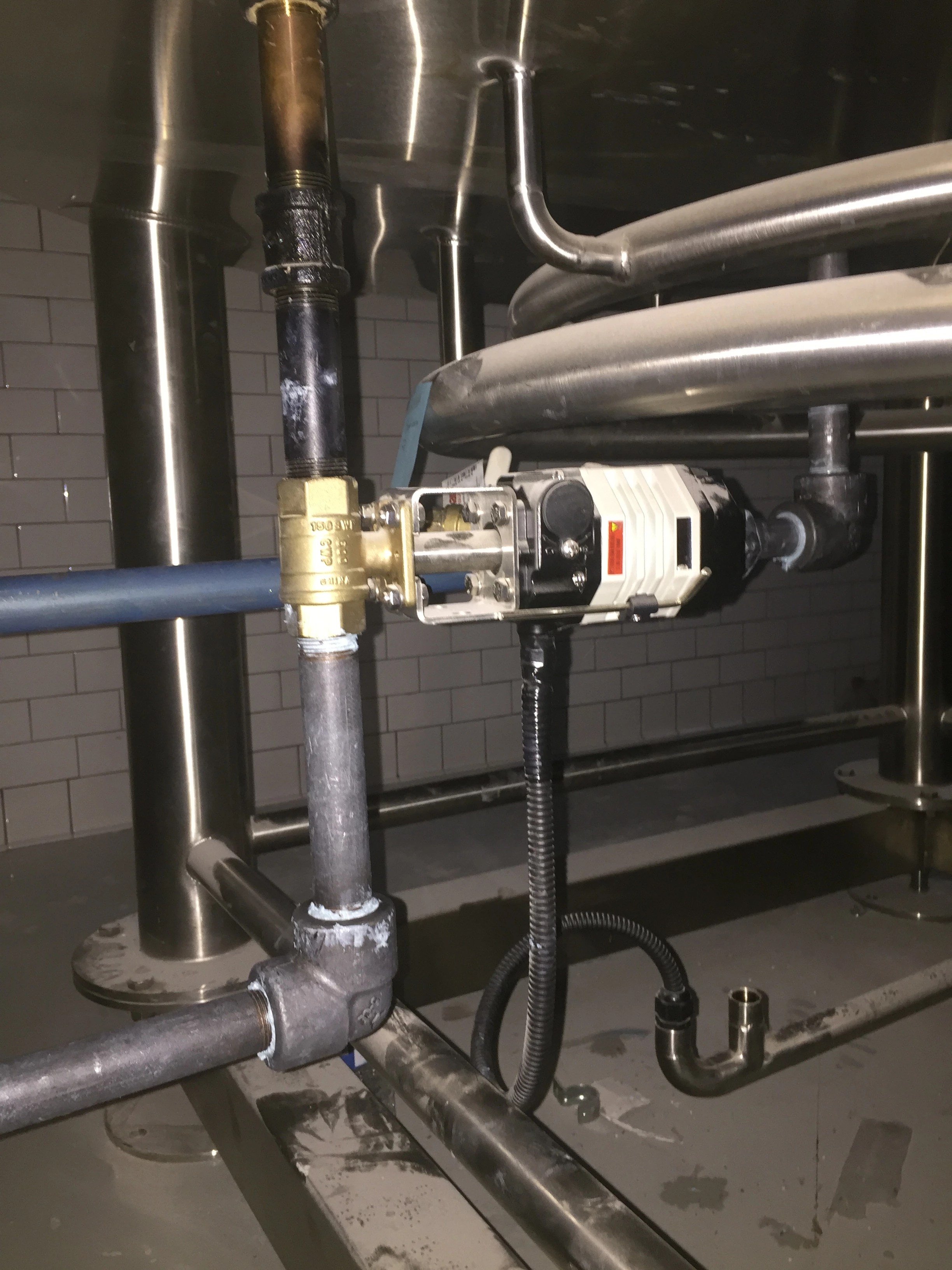 Electrically actuated brass ball valve with flexible conduit on utility piping beneath a stainless brewhouse vessel