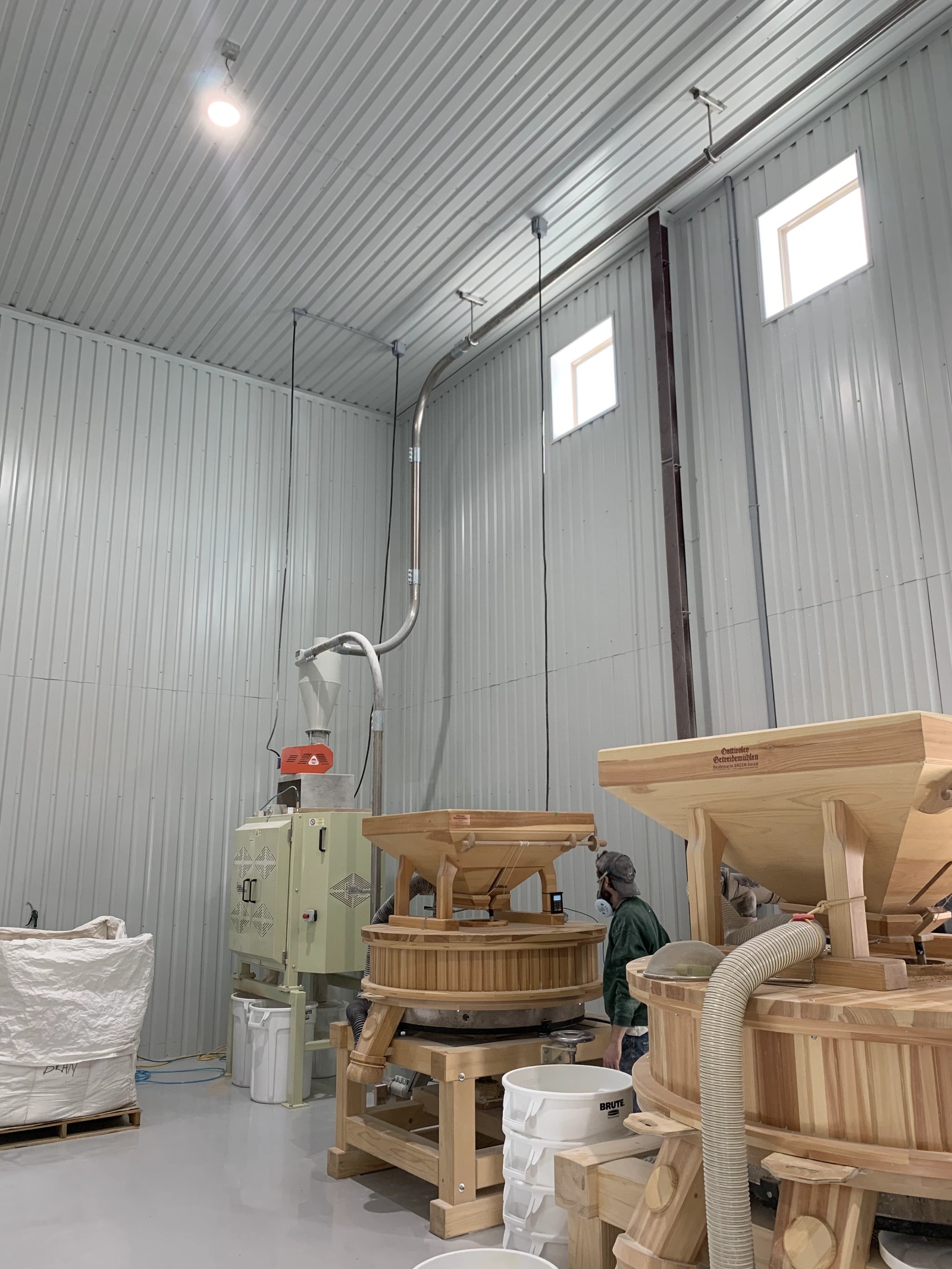 Two wooden stone grain mills, a cyclone collector, tote bags and a worker in a mill room