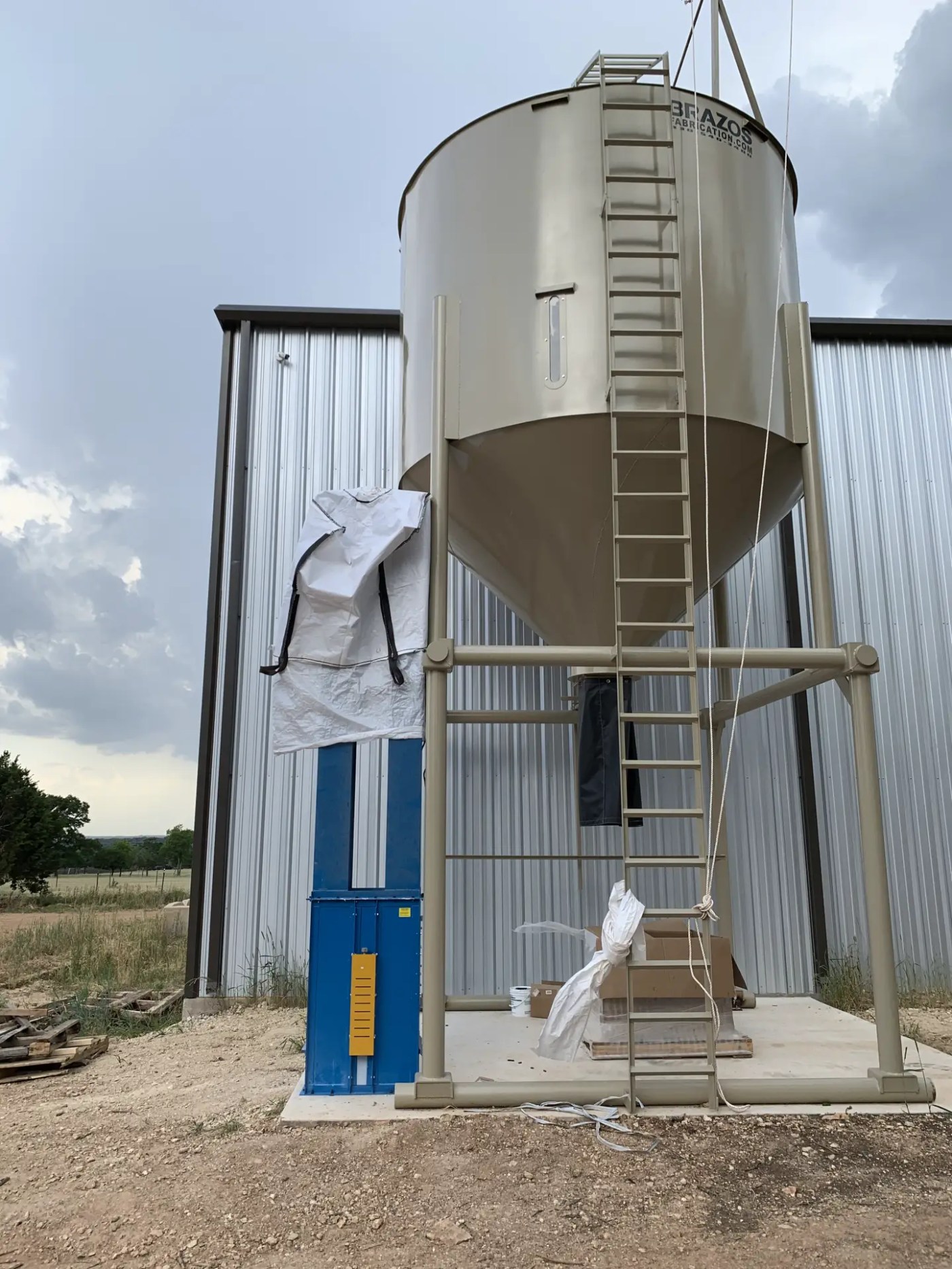 Elevated grain silo on a steel leg frame with a ladder, beside a metal building