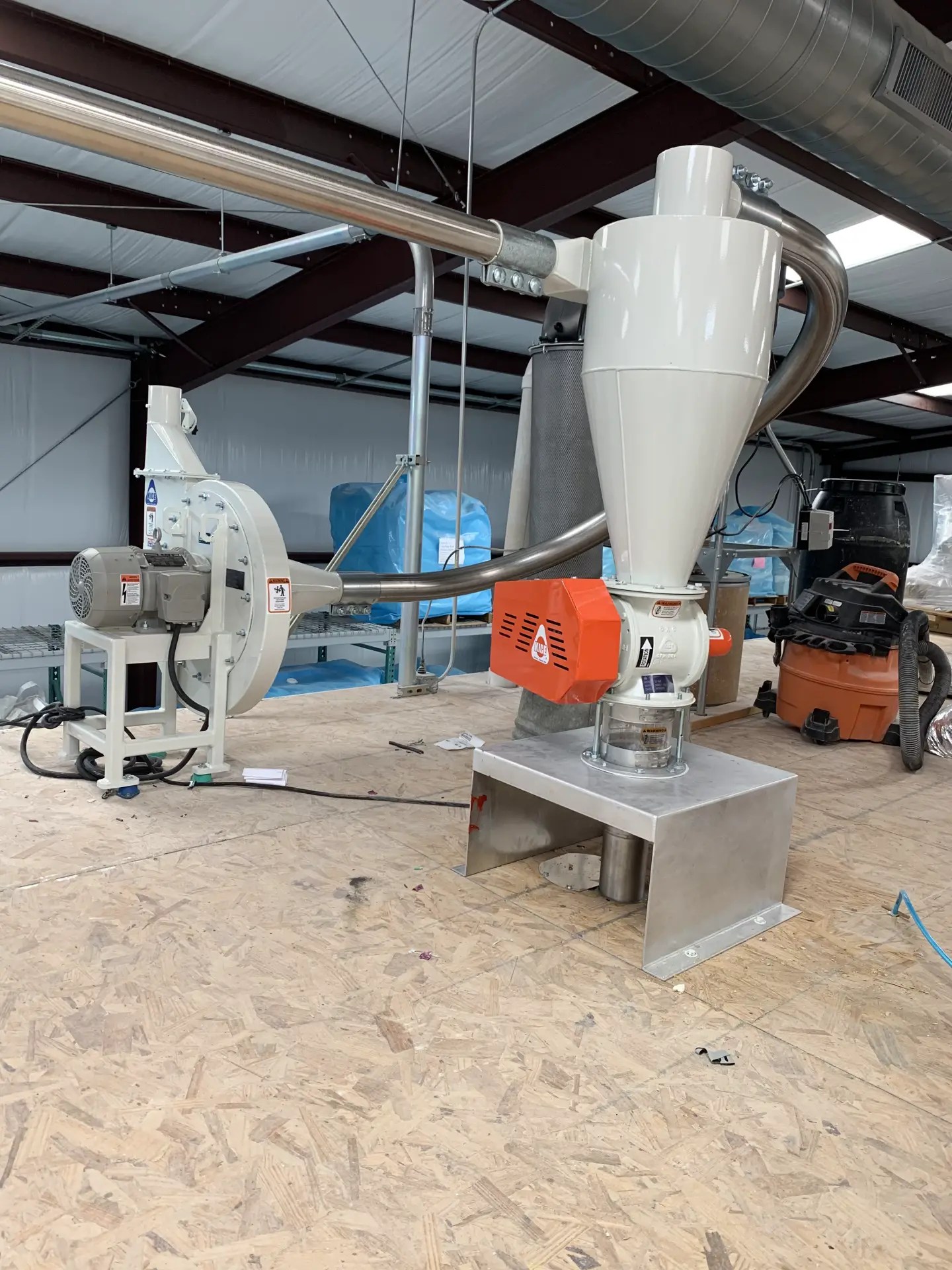 Motor-driven conveying blower and cyclone receiver linked by stainless pipe on a plywood warehouse mezzanine.