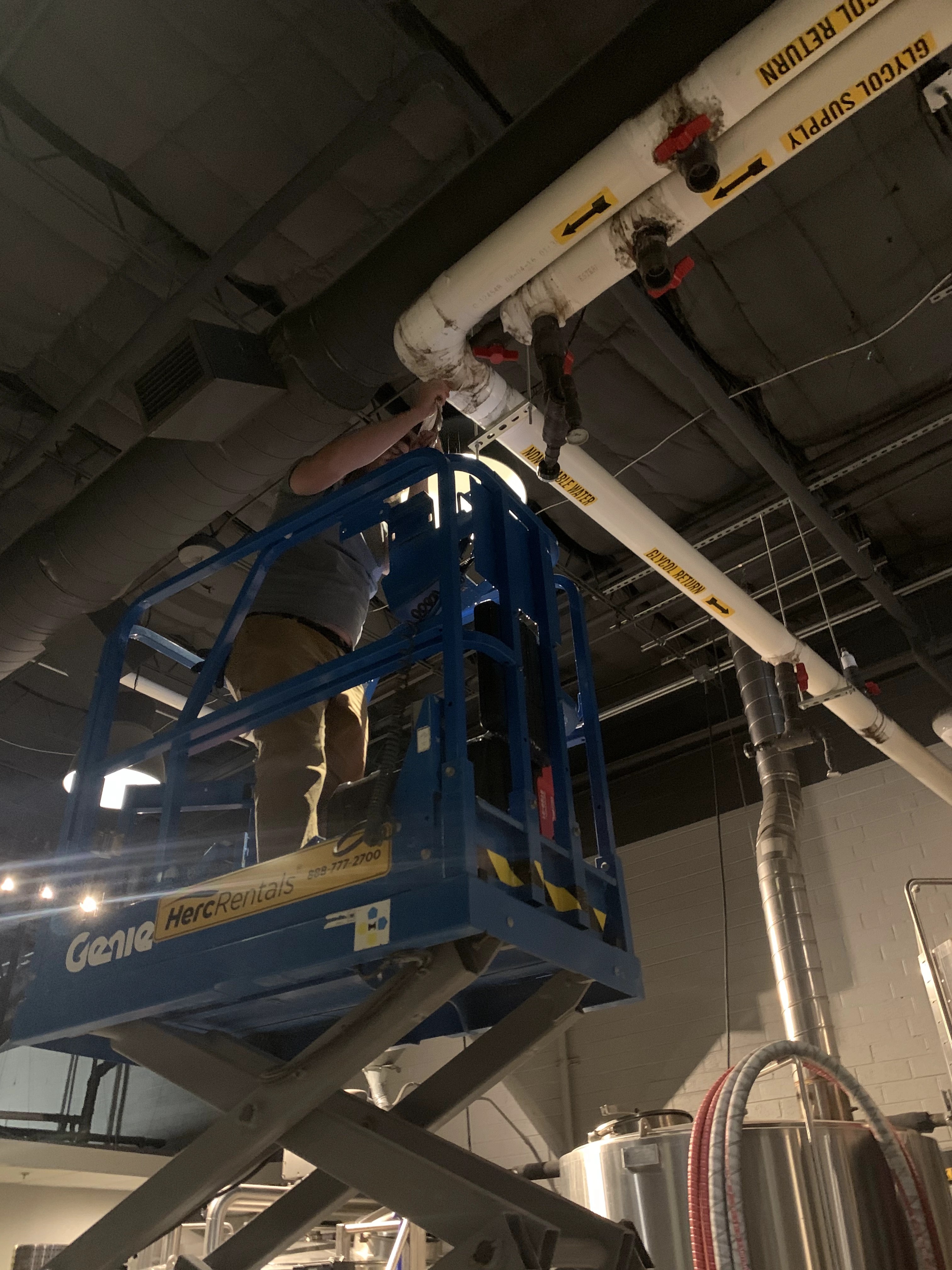 Worker on a scissor lift servicing labeled glycol supply and return piping above stainless tanks