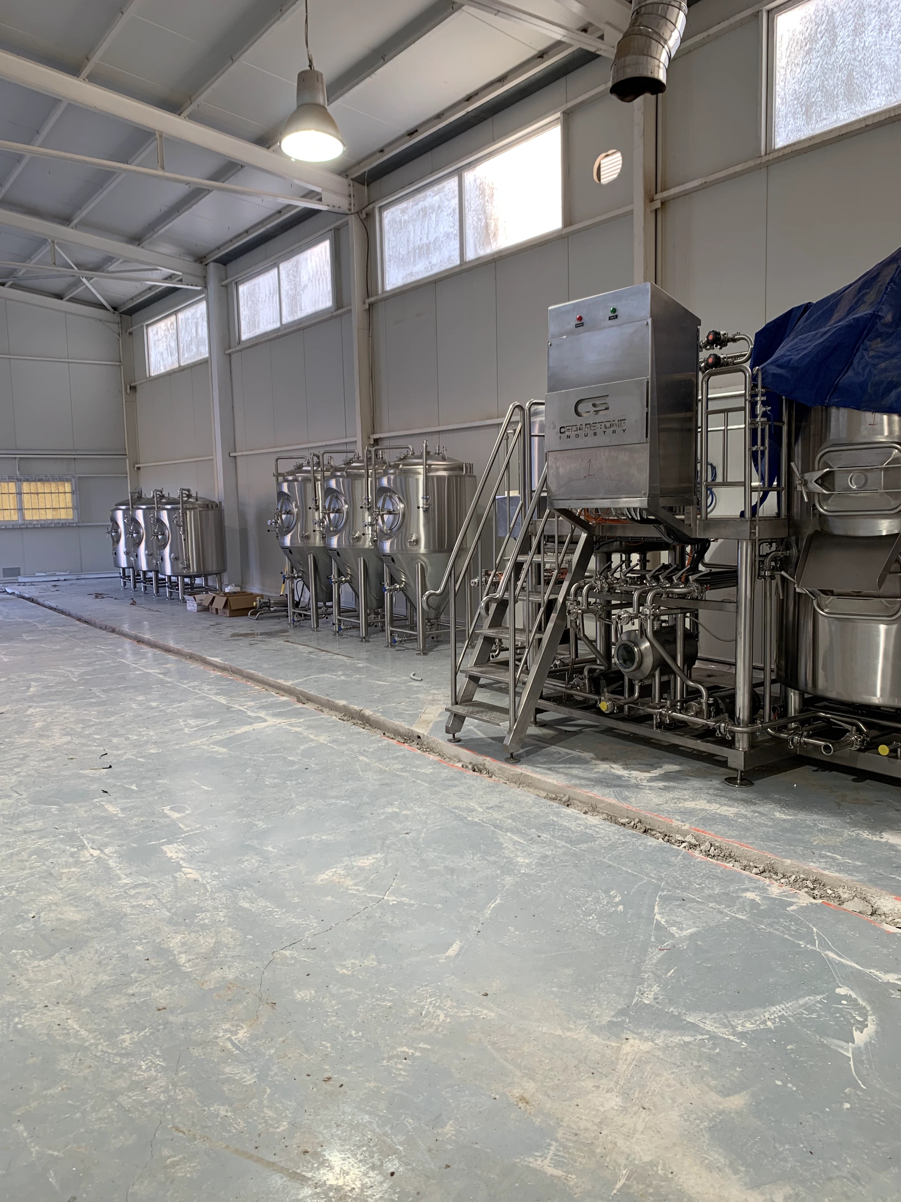 Newly placed process skid with platform stairs beside a row of fermenters on an unfinished floor.