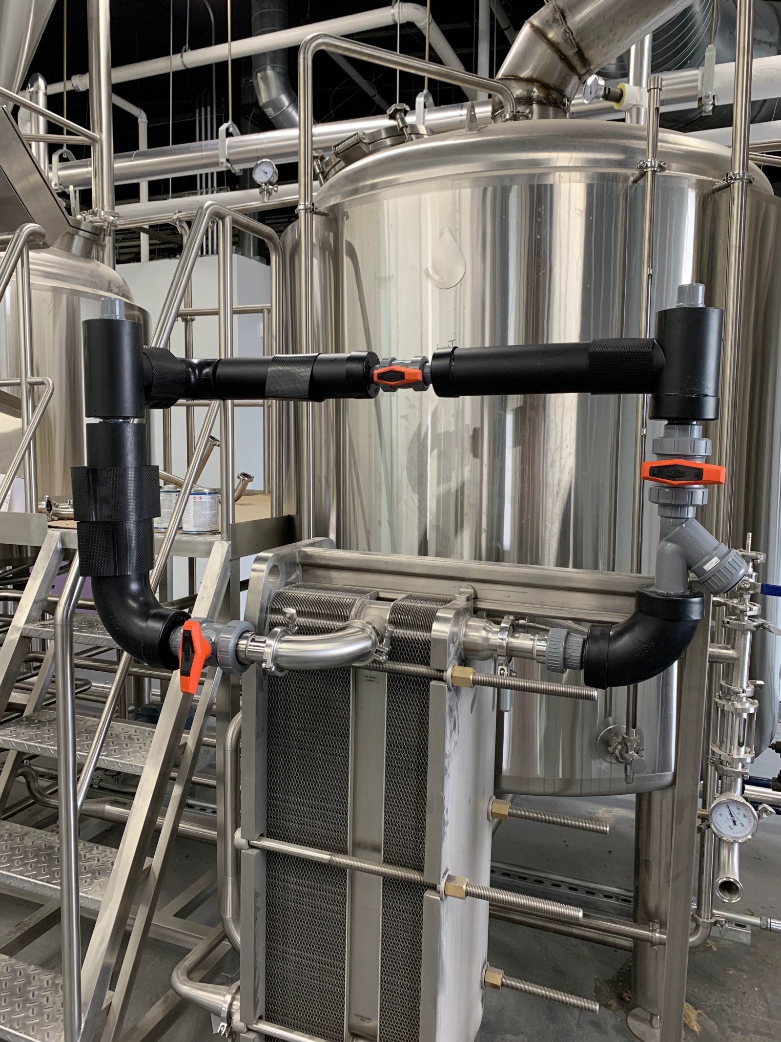 Plate heat exchanger with black piping and valves beside stainless brewhouse vessels