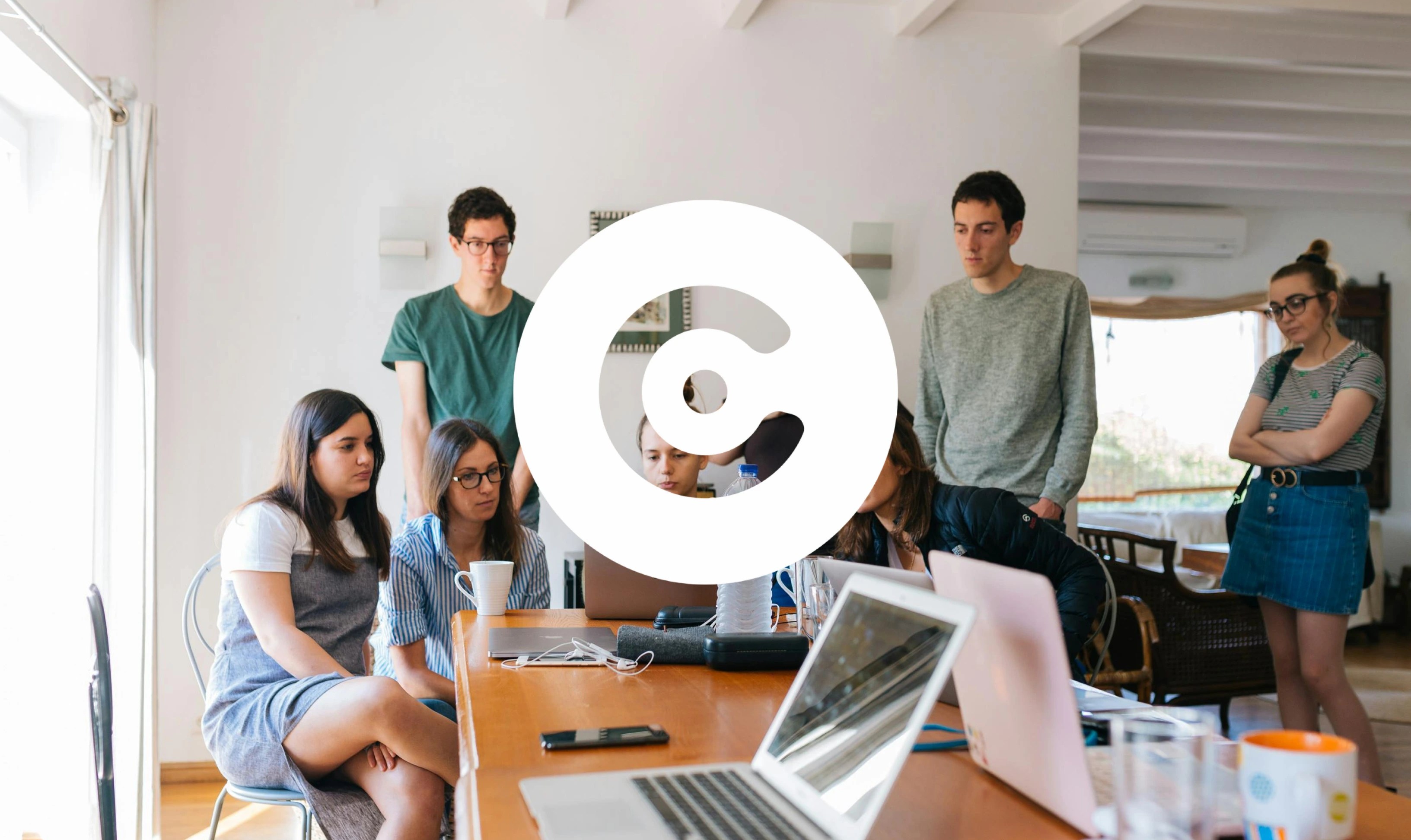 A group of people gathered around a table with laptops, looking at a shared screen or device during a collaborative work session, with a large circular logo overlaid in the center.