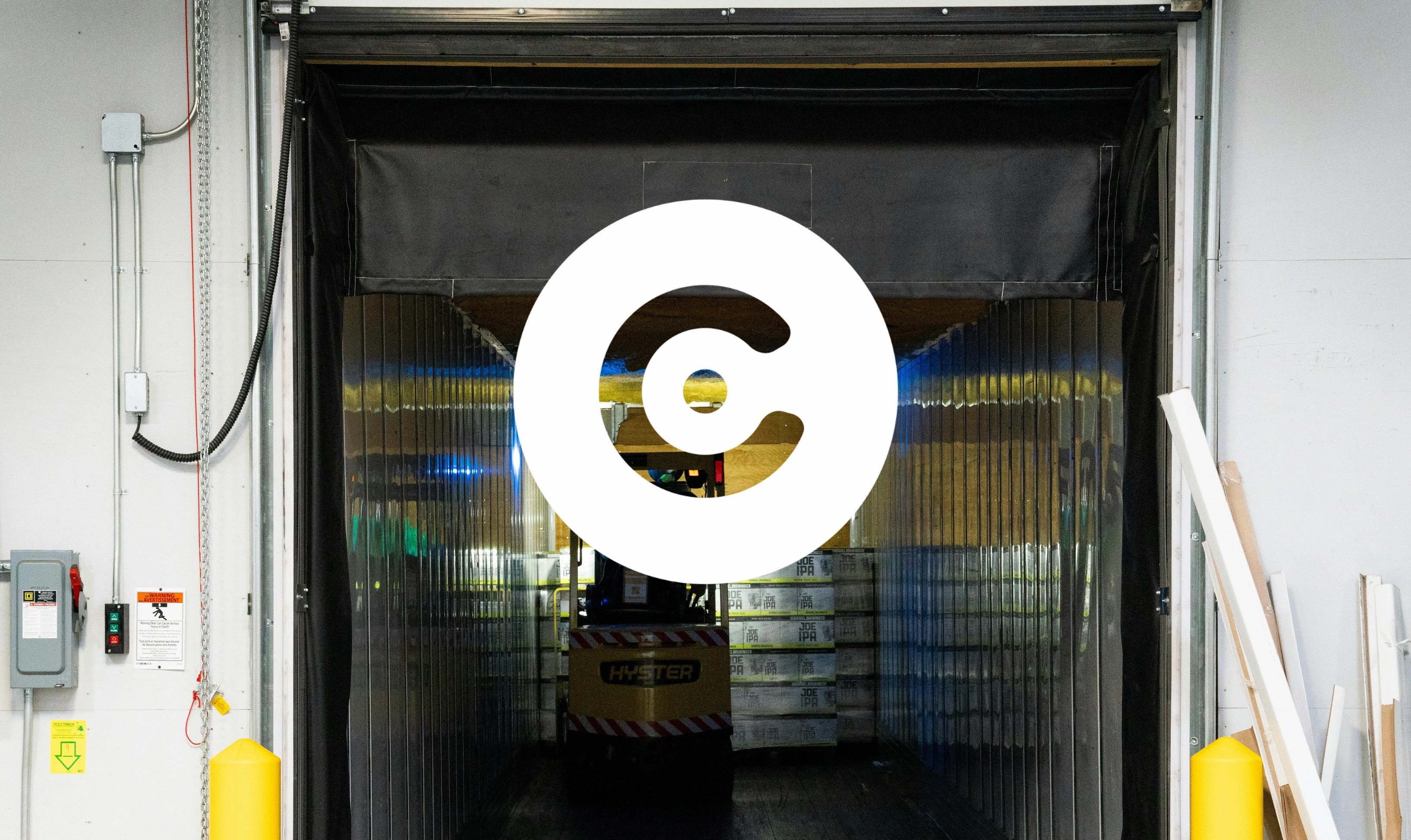 Forklift inside a warehouse loading dock, seen through transparent plastic strip curtains, with stacked pallets in the background and a large circular logo overlay in the center.