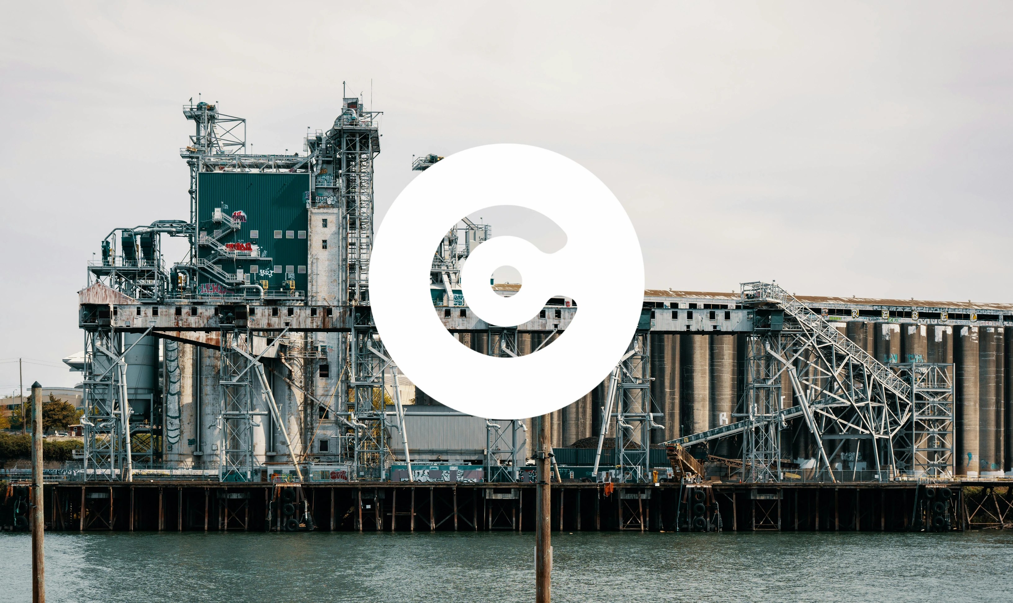 The image shows an industrial waterfront facility with large silos, metal frameworks, and conveyor structures. The buildings and equipment appear weathered, with some visible graffiti. The facility is positioned along the water, with structural supports extending into it. A large white circular logo with a central dot and cutout design is overlaid prominently in the center of the image.