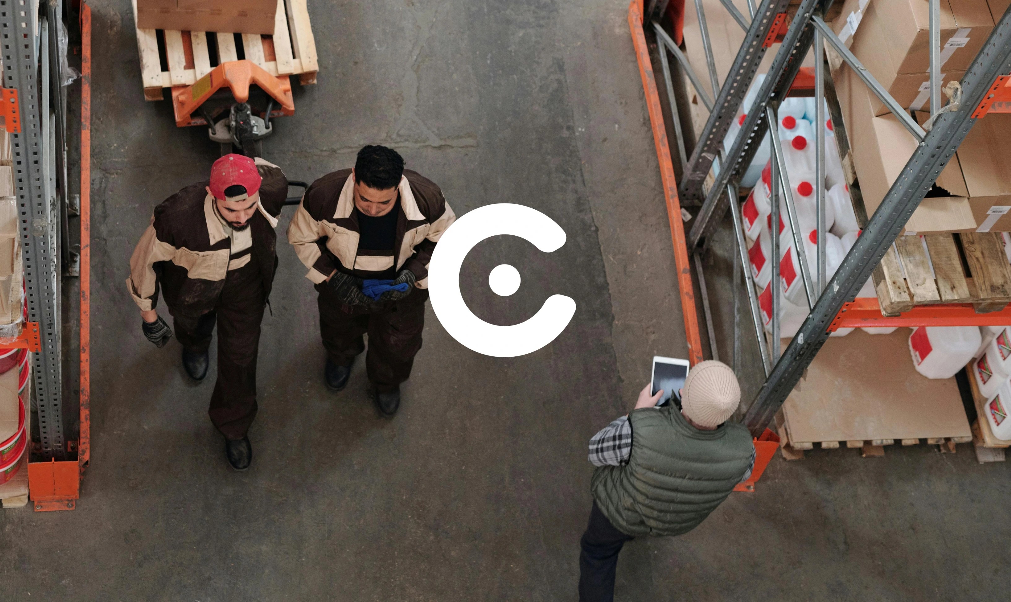 The image shows a warehouse setting with shelves stacked with boxes and containers. Two workers in brown uniforms walk down the aisle, one wearing a red cap. Another person, wearing a beanie and vest, holds a tablet and appears to be managing or checking inventory. A large white circular logo with a central dot and cutout design is overlaid prominently in the center of the image.