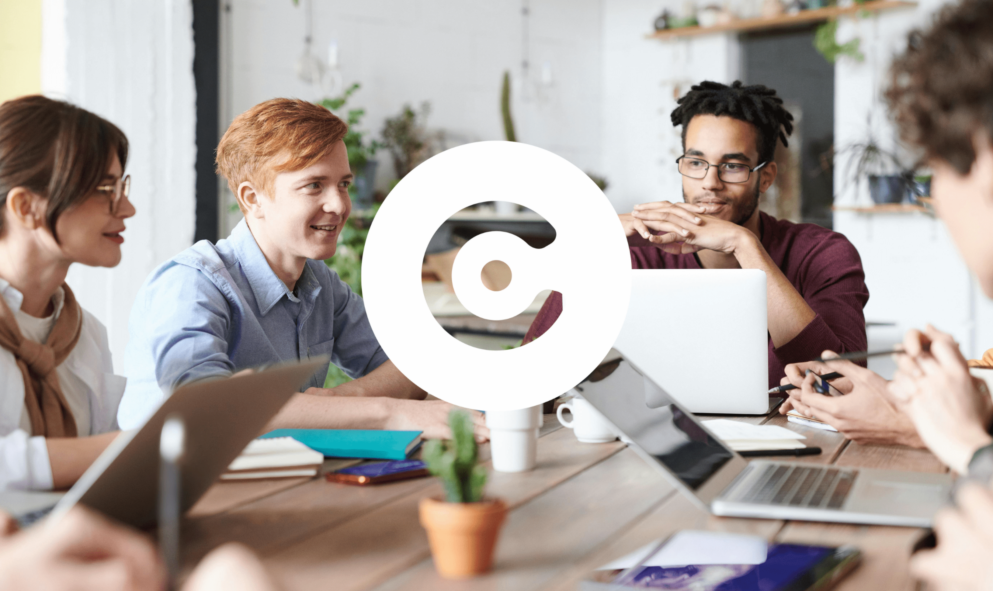 Group of people sitting around a table with laptops and notebooks, having a discussion in a modern office space. A large white circular logo is overlaid in the center.