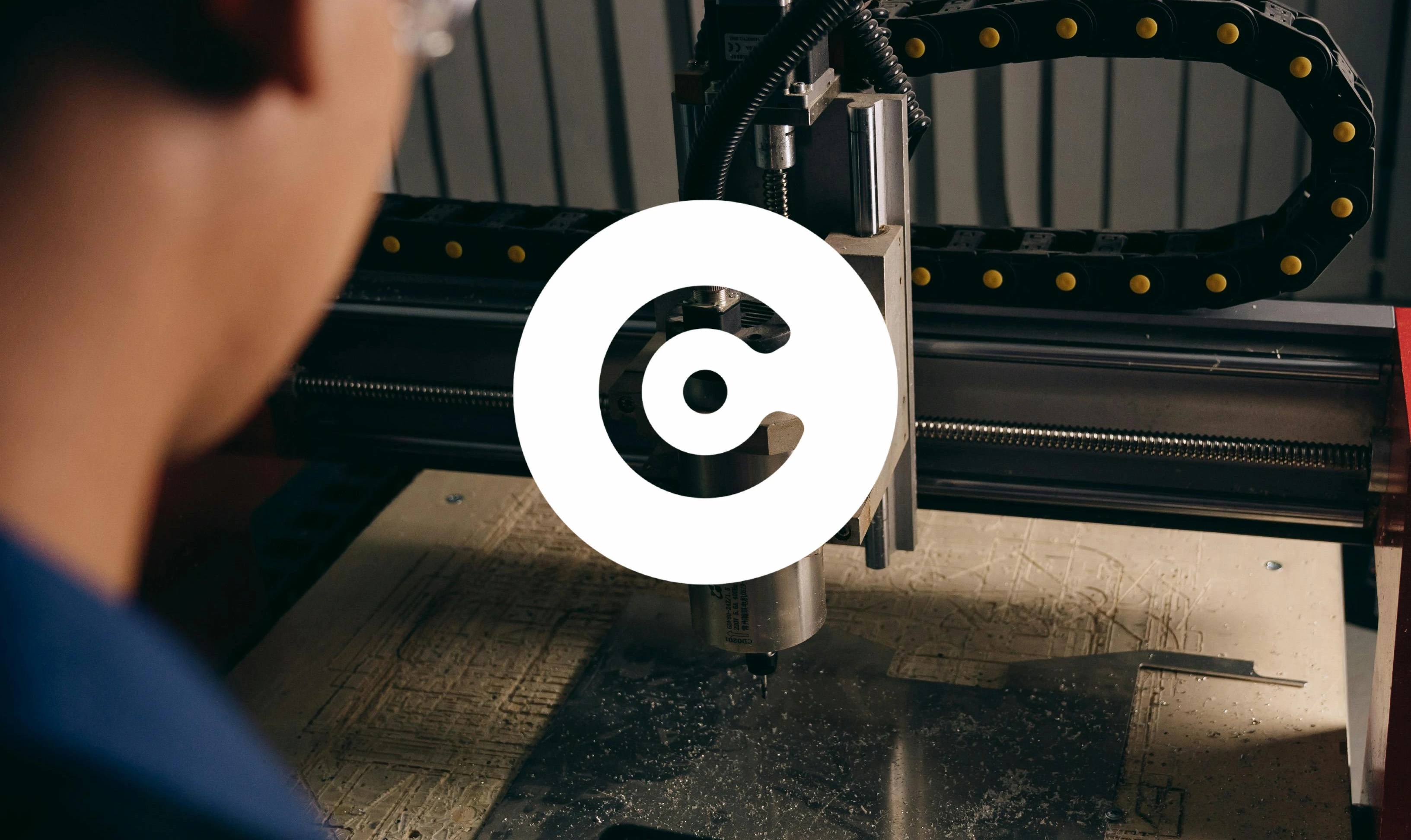 The image shows a person operating a CNC (Computer Numerical Control) machine, which is actively engraving or cutting a material -likely wood or plastic - on a flat surface. The foreground is blurred, focusing on the machine’s spindle and cutting head. A large white circular logo with a dot and curved line overlay is placed at the center of the image.