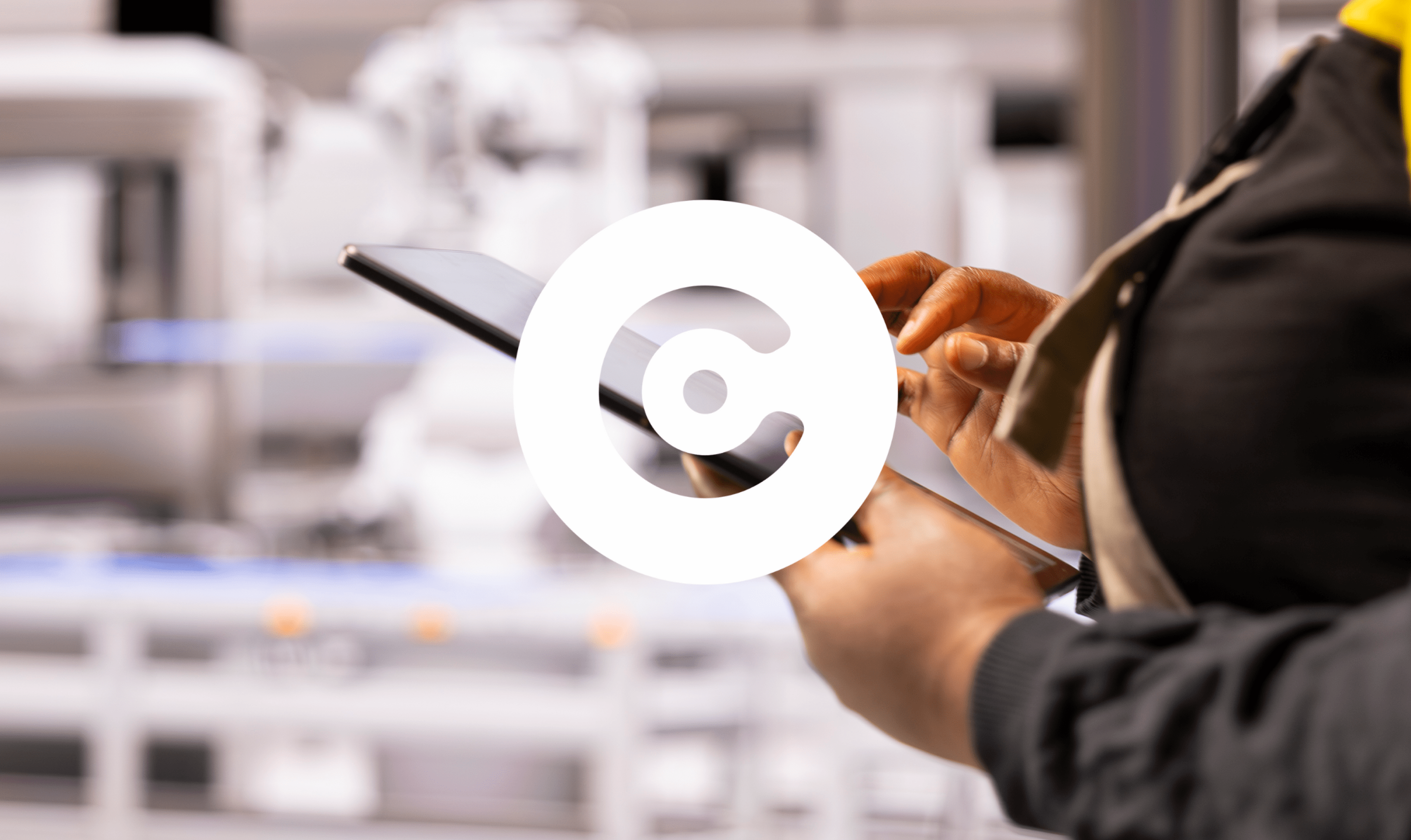 Close-up of a person using a tablet in an industrial or factory setting, with blurred machinery in the background. A large white circular logo is overlaid in the center.