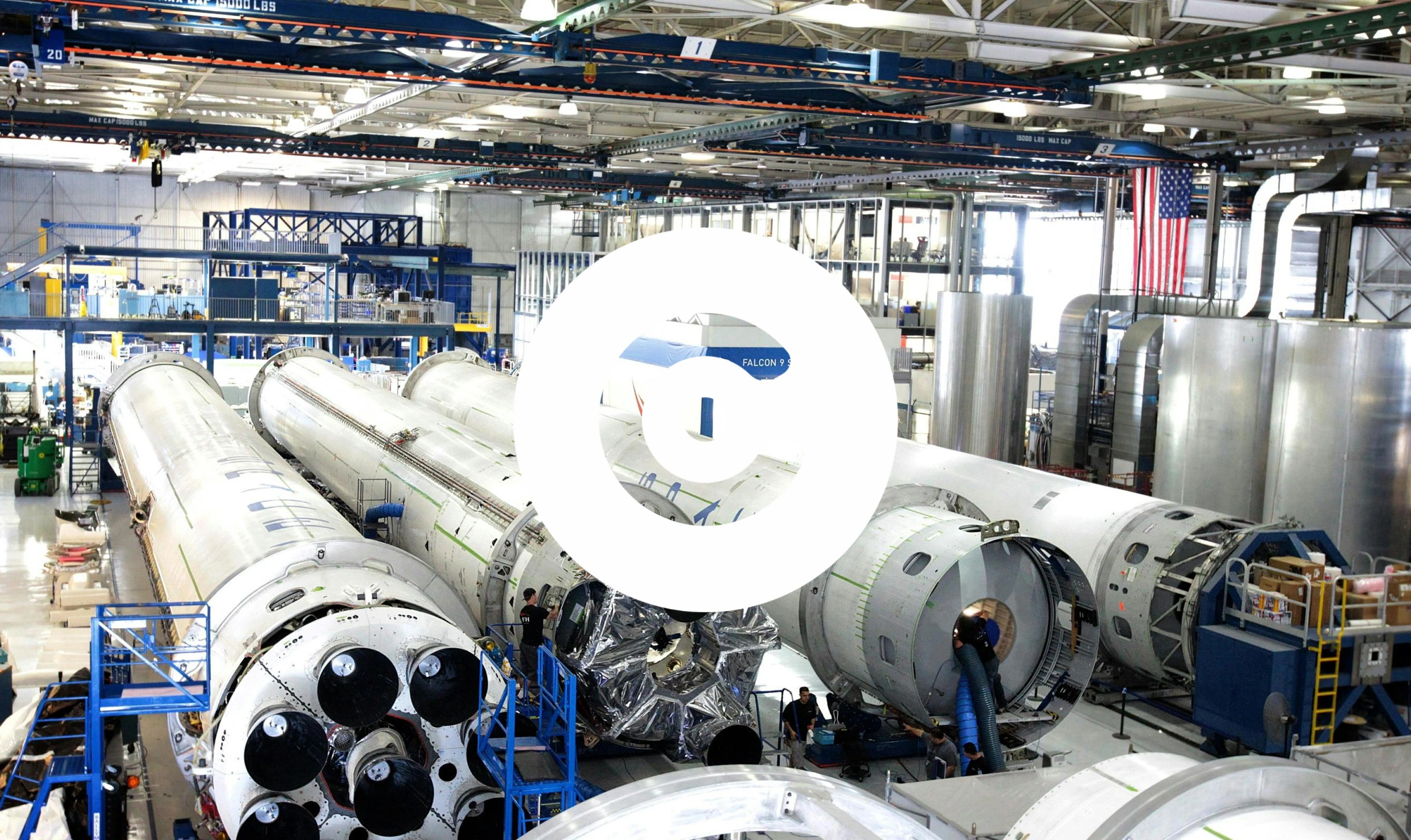 The image shows the interior of a large aerospace manufacturing facility with several rocket boosters being assembled or serviced. Engineers and technicians can be seen working around the rockets, with scaffolding, equipment, and cranes in the background. The U.S. flag hangs on the right side, and the facility is brightly lit with high ceilings and industrial machinery.