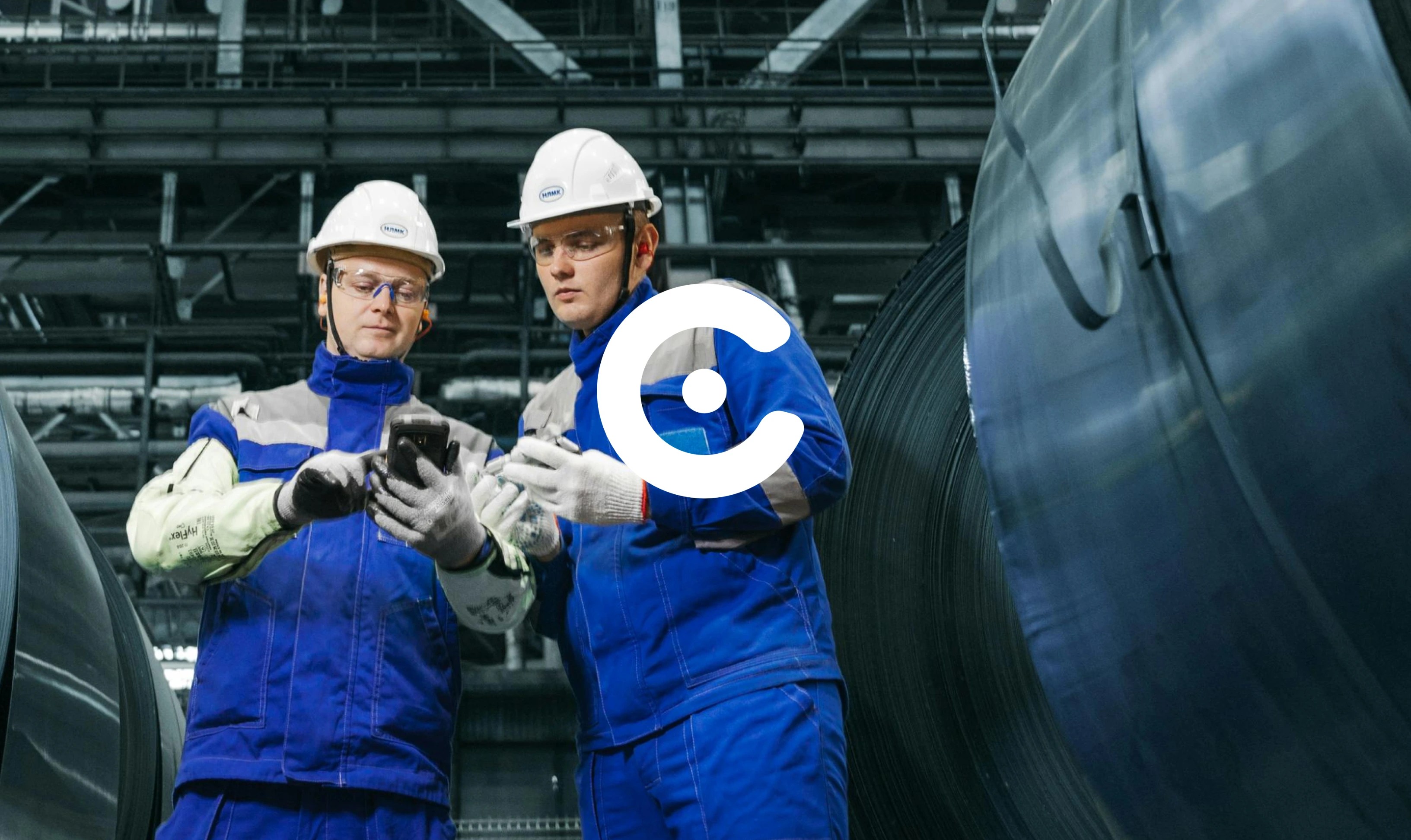 Two industrial workers in blue uniforms and white helmets examine a smartphone in a factory setting, standing between large metal coils. A white circular logo is overlaid at the center of the image.