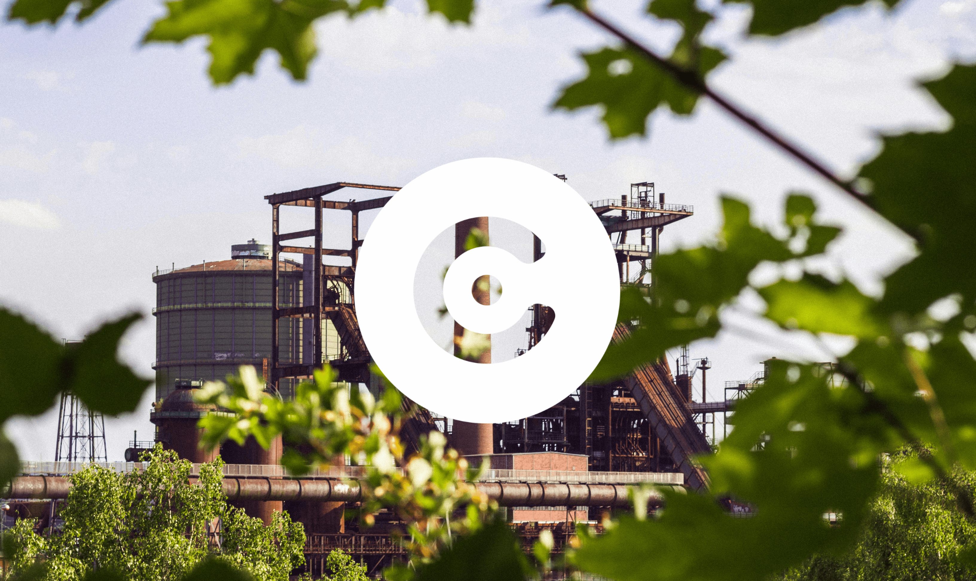 View of a large industrial facility with tanks, pipes, and steel structures, framed by green tree leaves in the foreground.