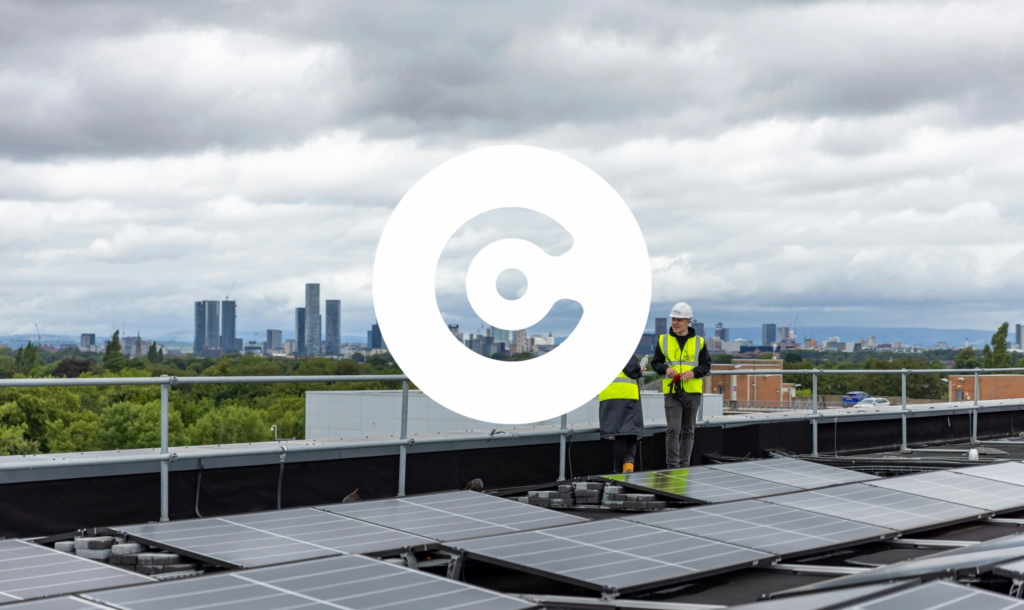 Solar panels on a rooftop with two workers in safety vests and helmets talking, with a city skyline under cloudy skies in the background.