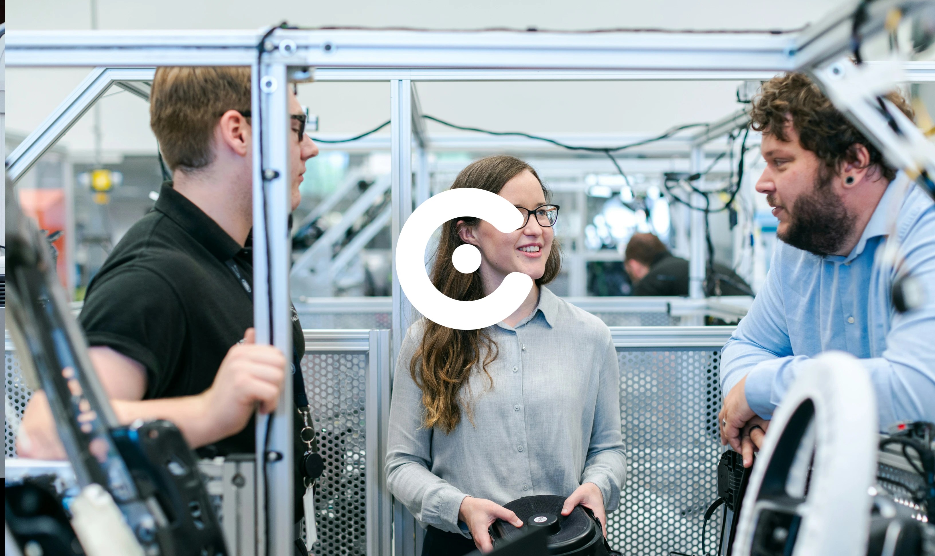 Three people standing and talking in a manufacturing or engineering workspace, surrounded by machinery and metal frames.