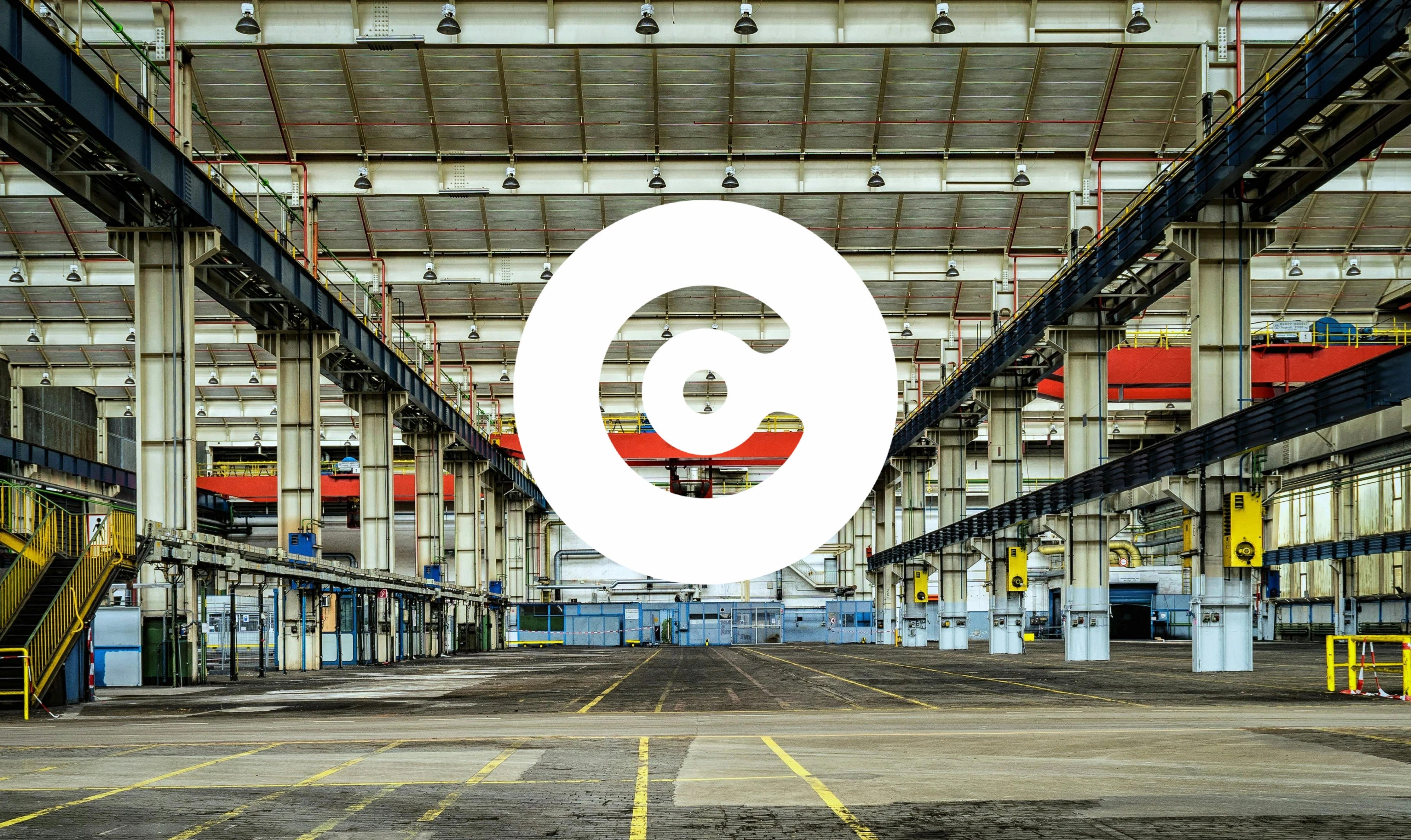 Wide view of an empty industrial warehouse with tall support columns, overhead lighting, and a large white circular logo centered in the foreground.