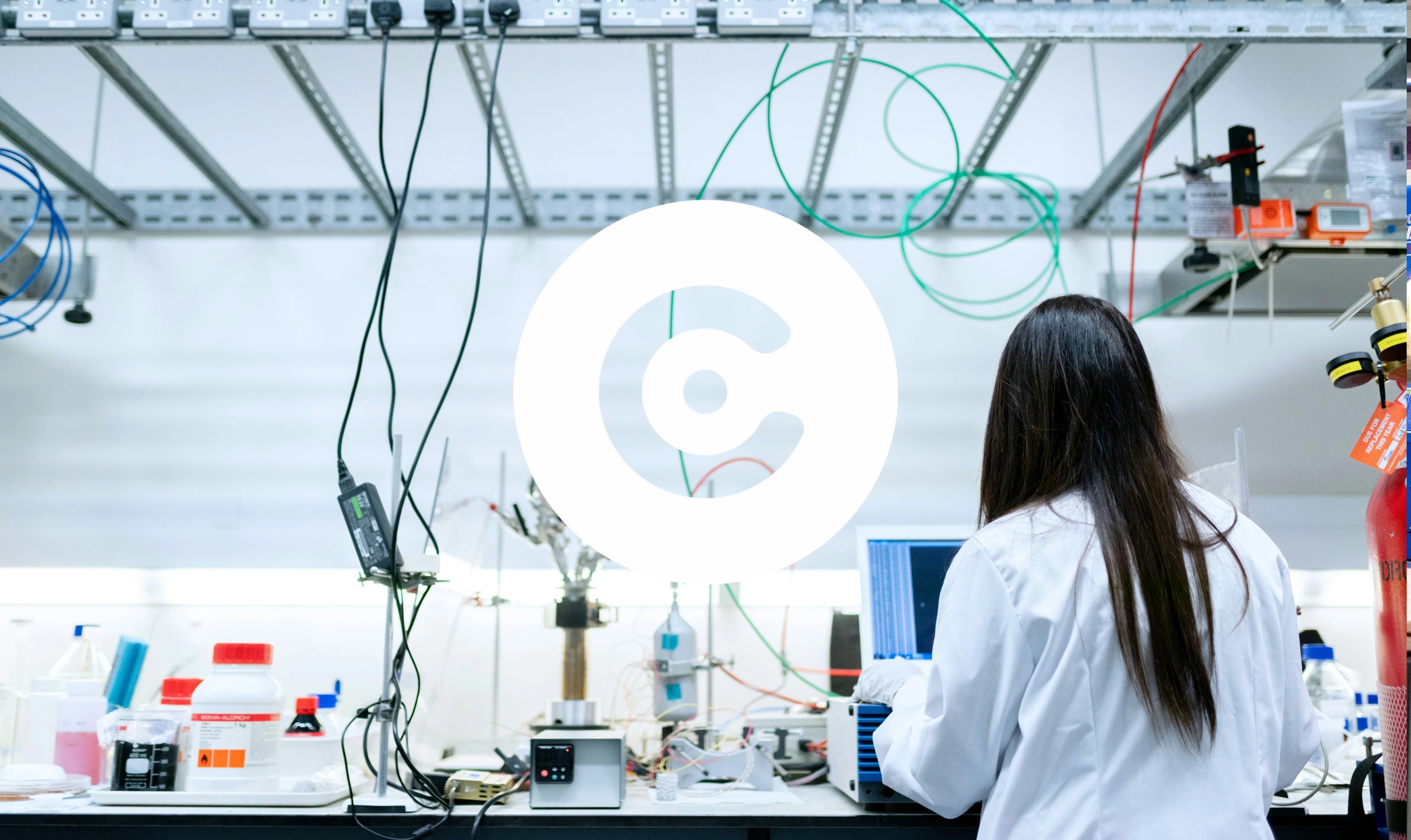 A person in a white lab coat works at a cluttered laboratory bench with scientific equipment, wires, and a laptop, surrounded by cables and containers in a research lab setting.