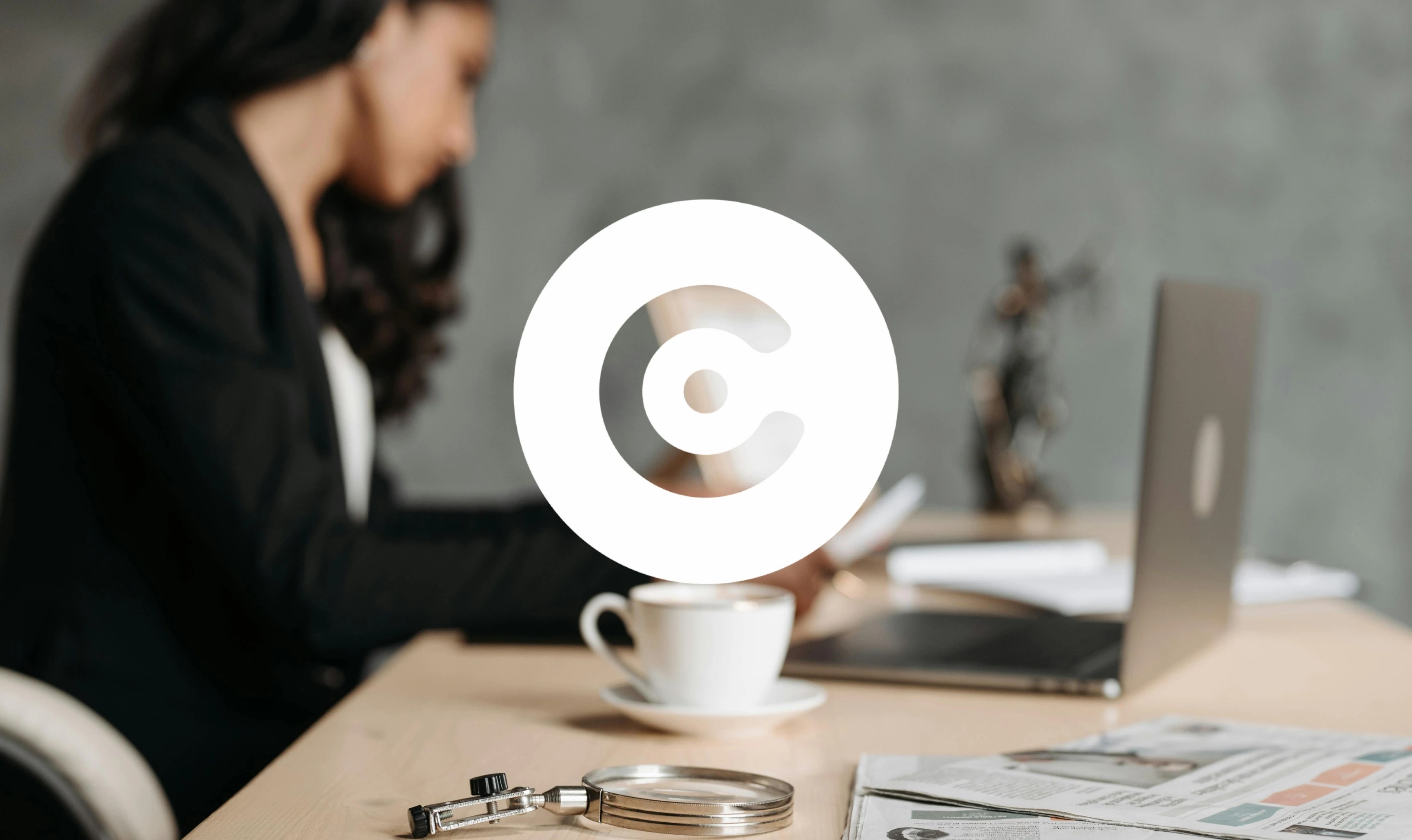 A blurred woman working at a desk with a laptop, coffee cup, and office tools in the foreground, overlaid with a large white circular logo.