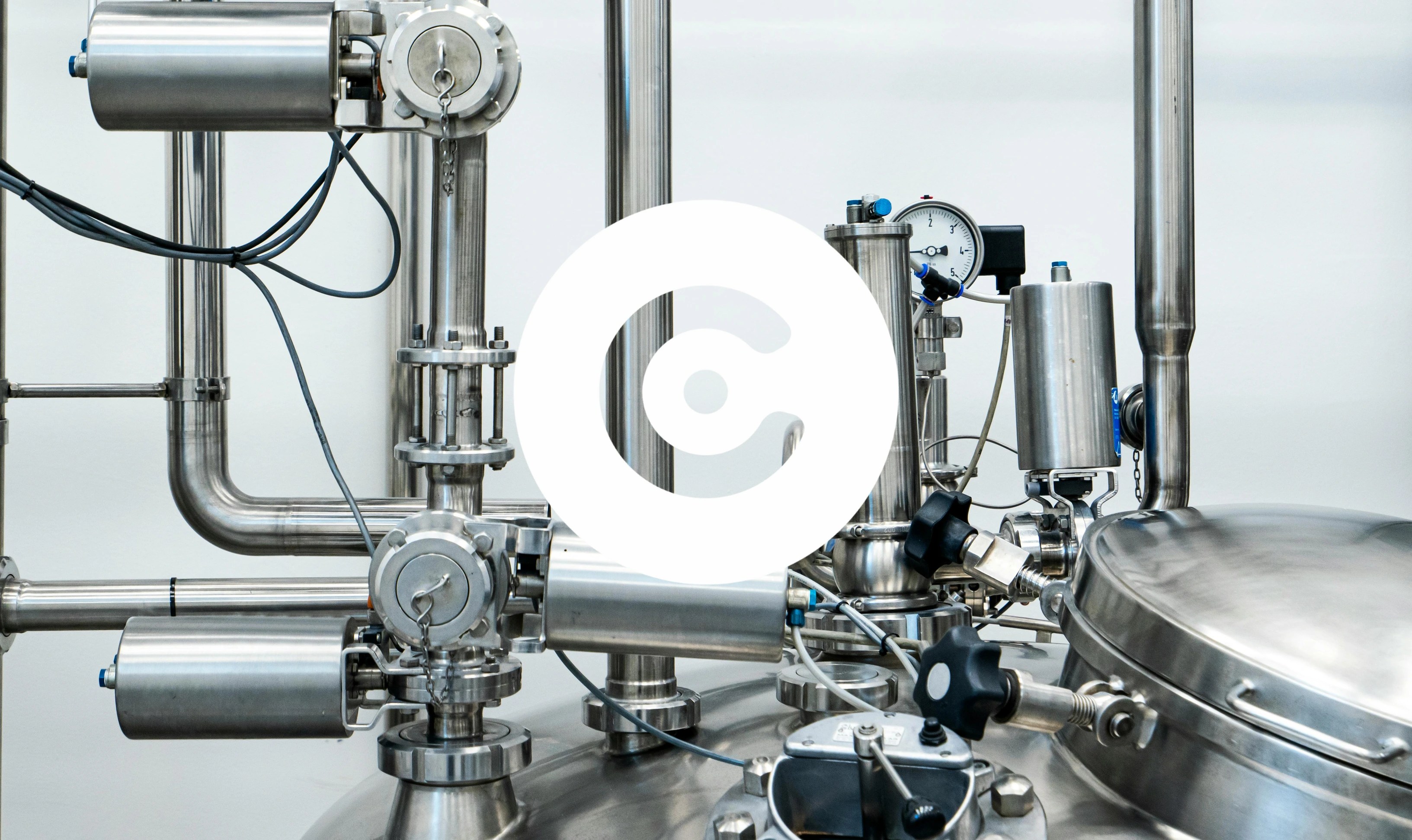 Close-up of stainless steel industrial machinery with pipes, valves, gauges and a large sealed tank in a clean manufacturing environment.