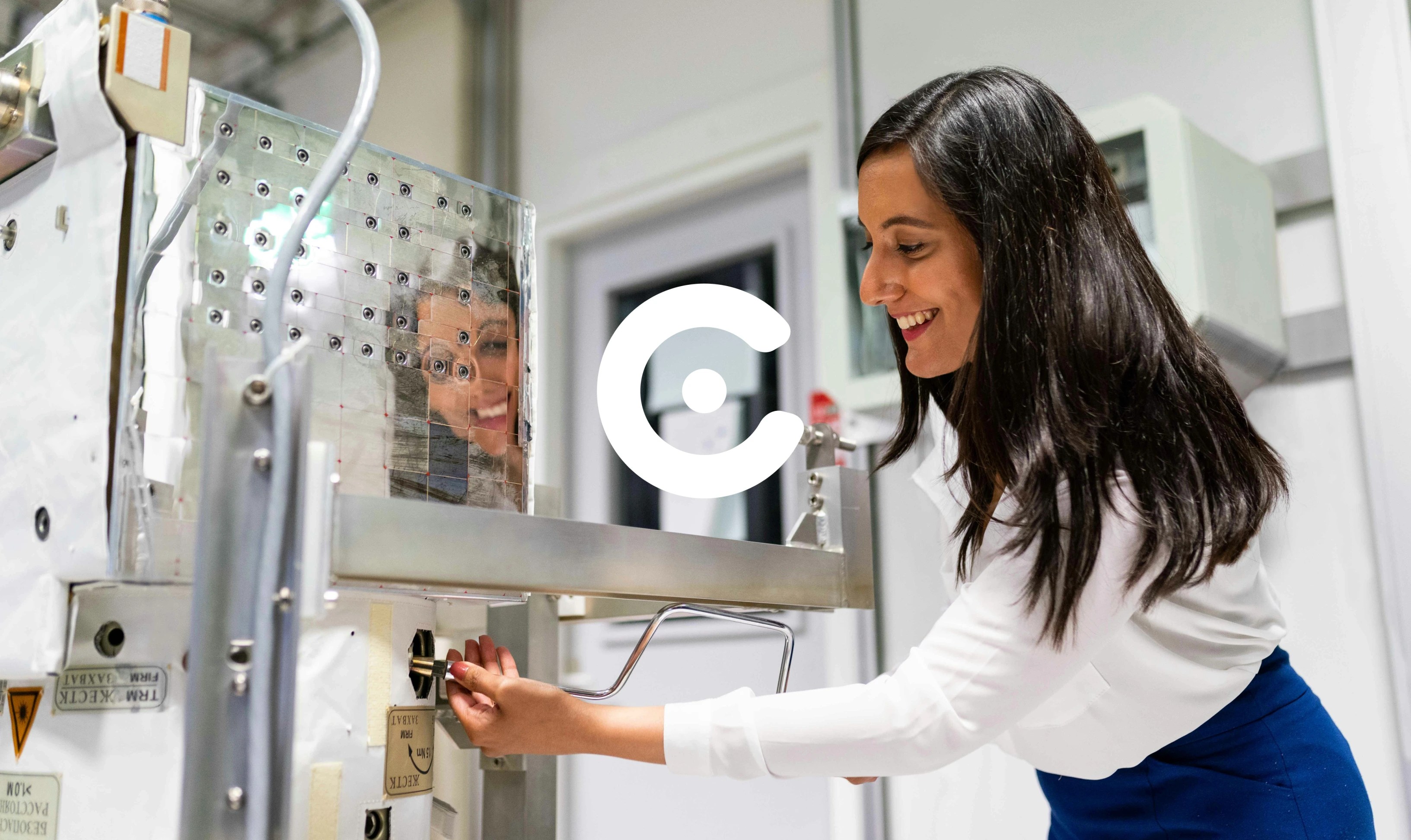 A smiling woman operates a piece of industrial equipment, with her reflection visible on a metallic panel in front of her.