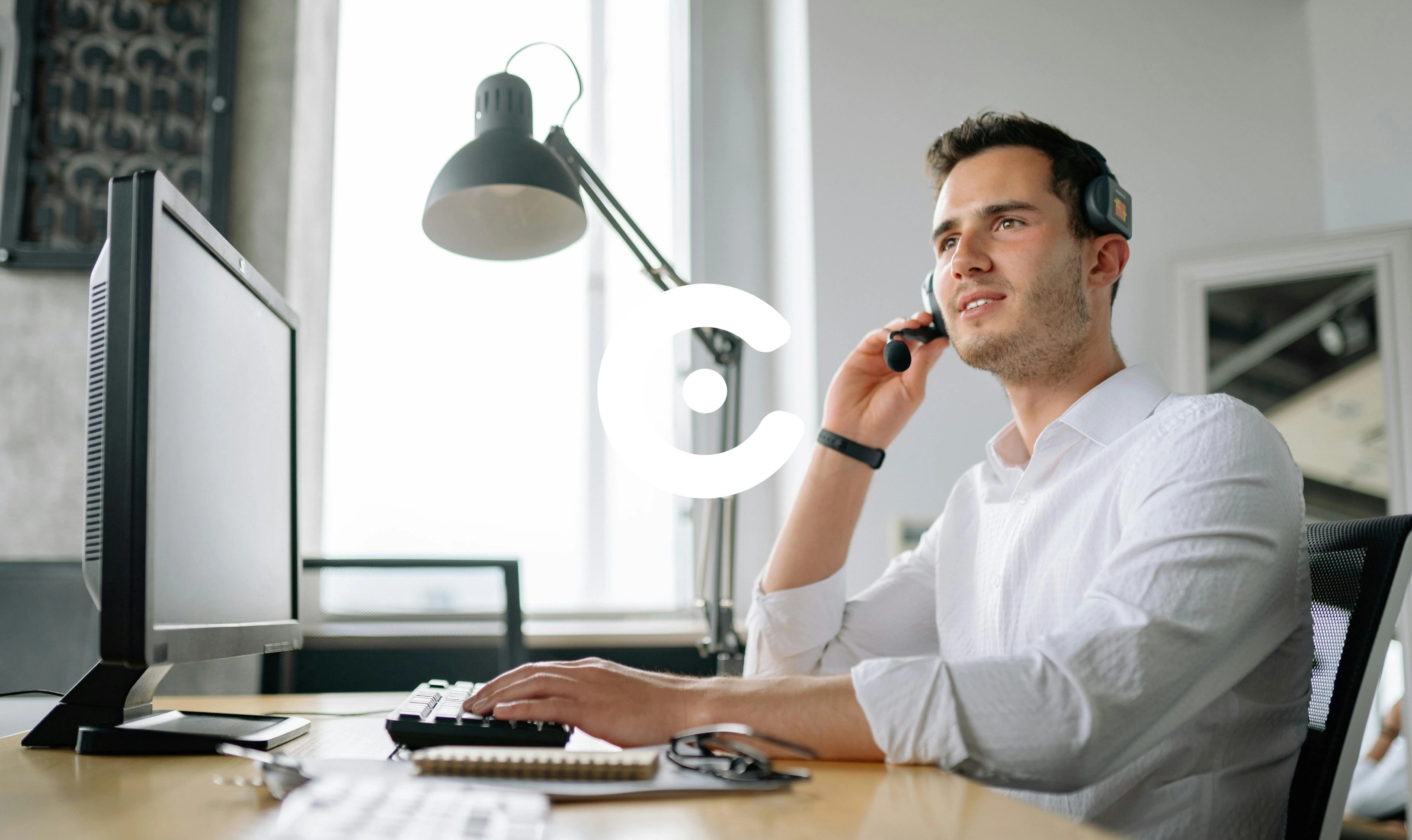 Man wearing a headset and white shirt sits at a desk, speaking into a microphone while working on a computer in a modern office.