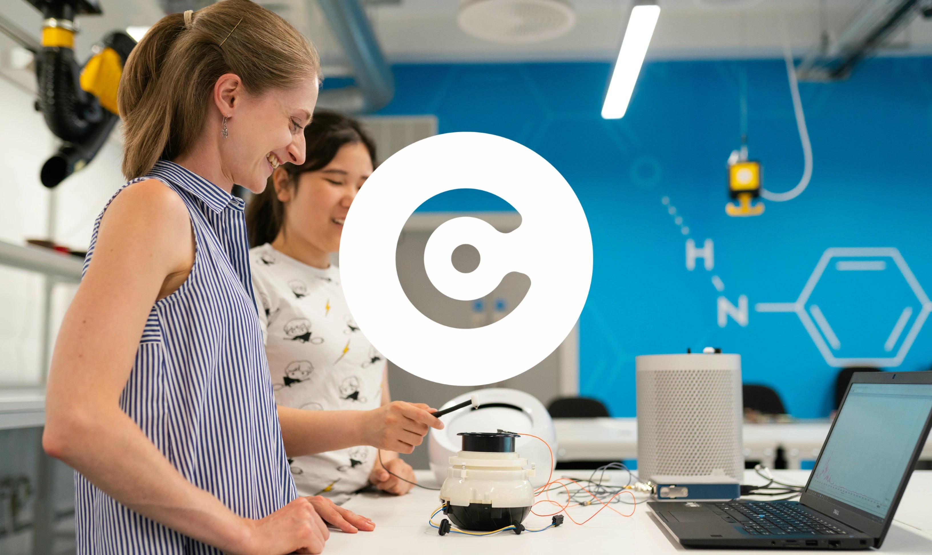 Two women in a modern lab workspace smile while testing an electronic prototype connected to a laptop on a worktable.