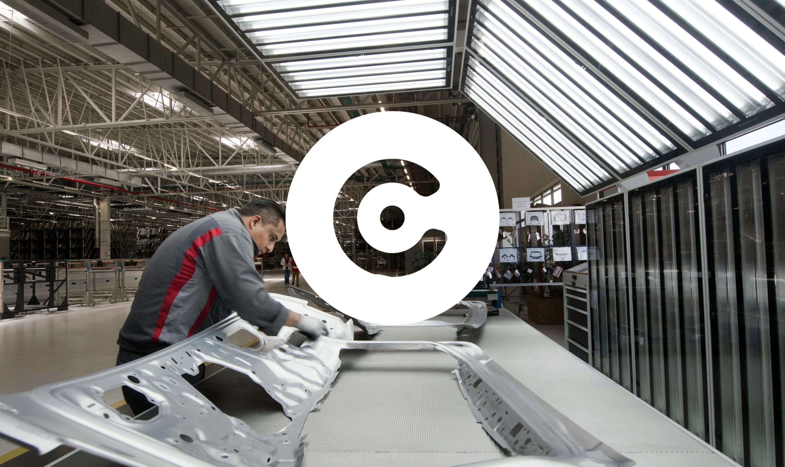 Factory worker inspecting or assembling large metal car body panels on a production line inside a modern automotive manufacturing plant.