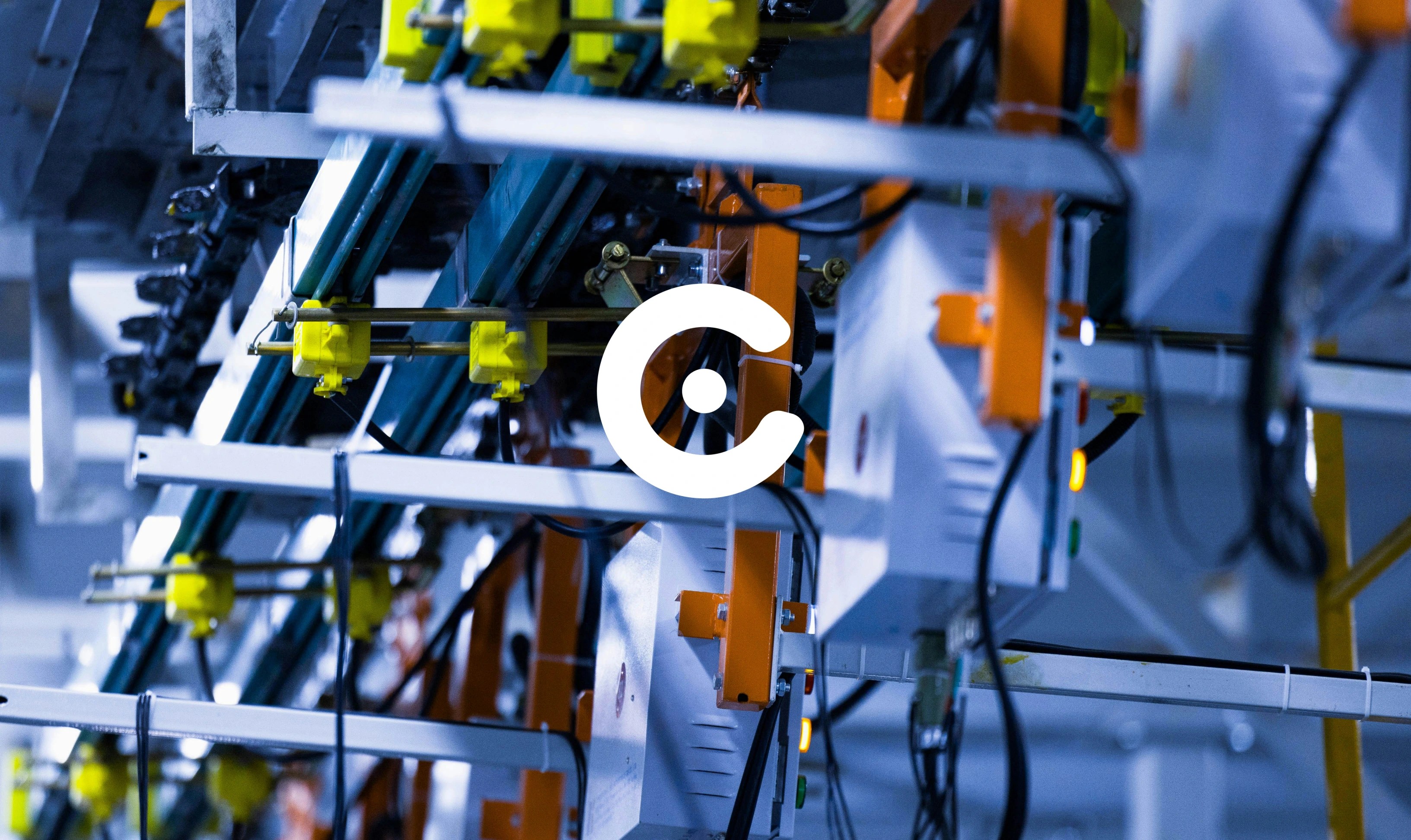 A white stylized letter "C" with a dot in the center is overlaid on a blue-toned industrial manufacturing assembly line with orange and yellow machinery components.