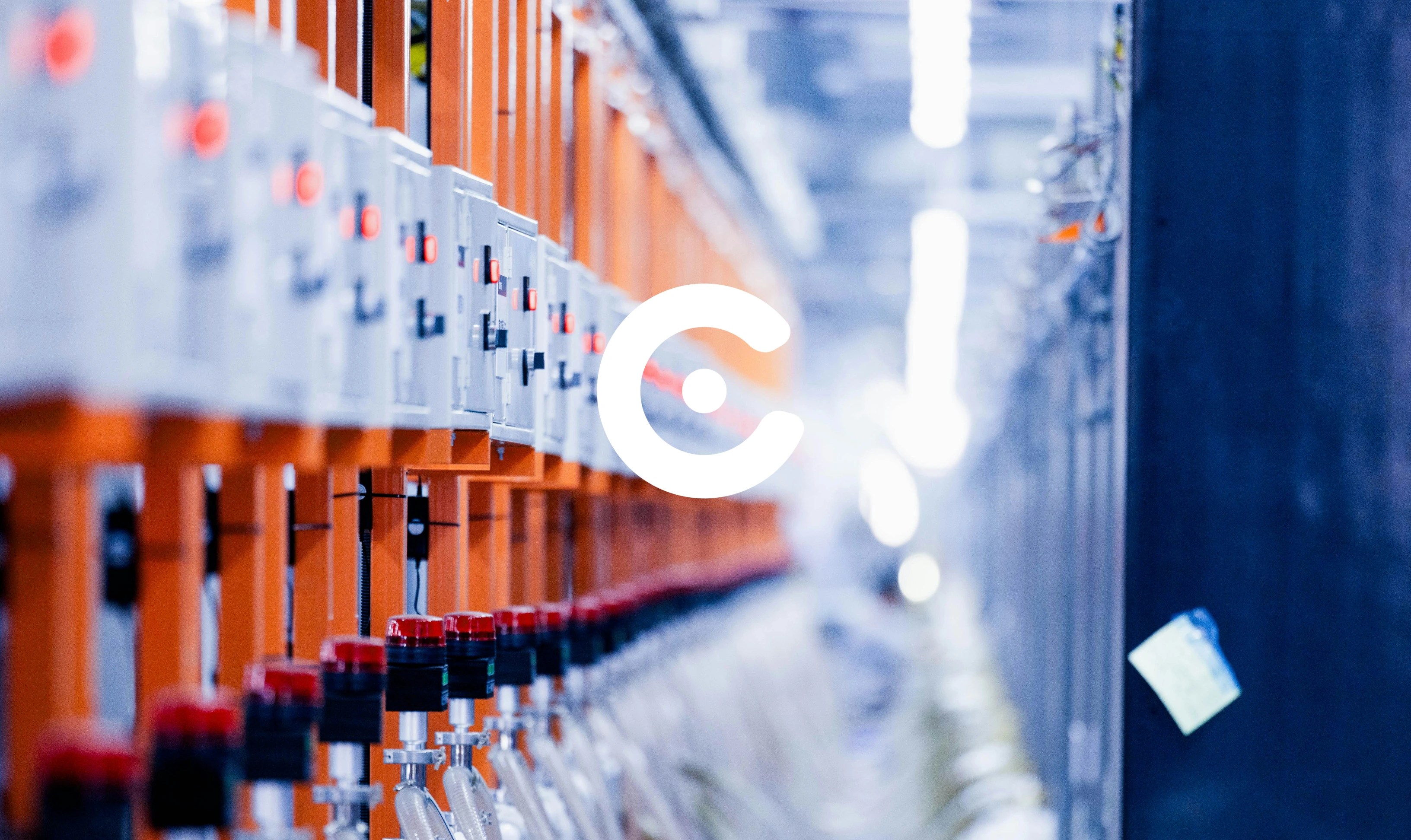 Row of industrial electrical control panels with switches and red indicator lights mounted on orange racks in a factory or technical facility, with cables and tubing running below.
