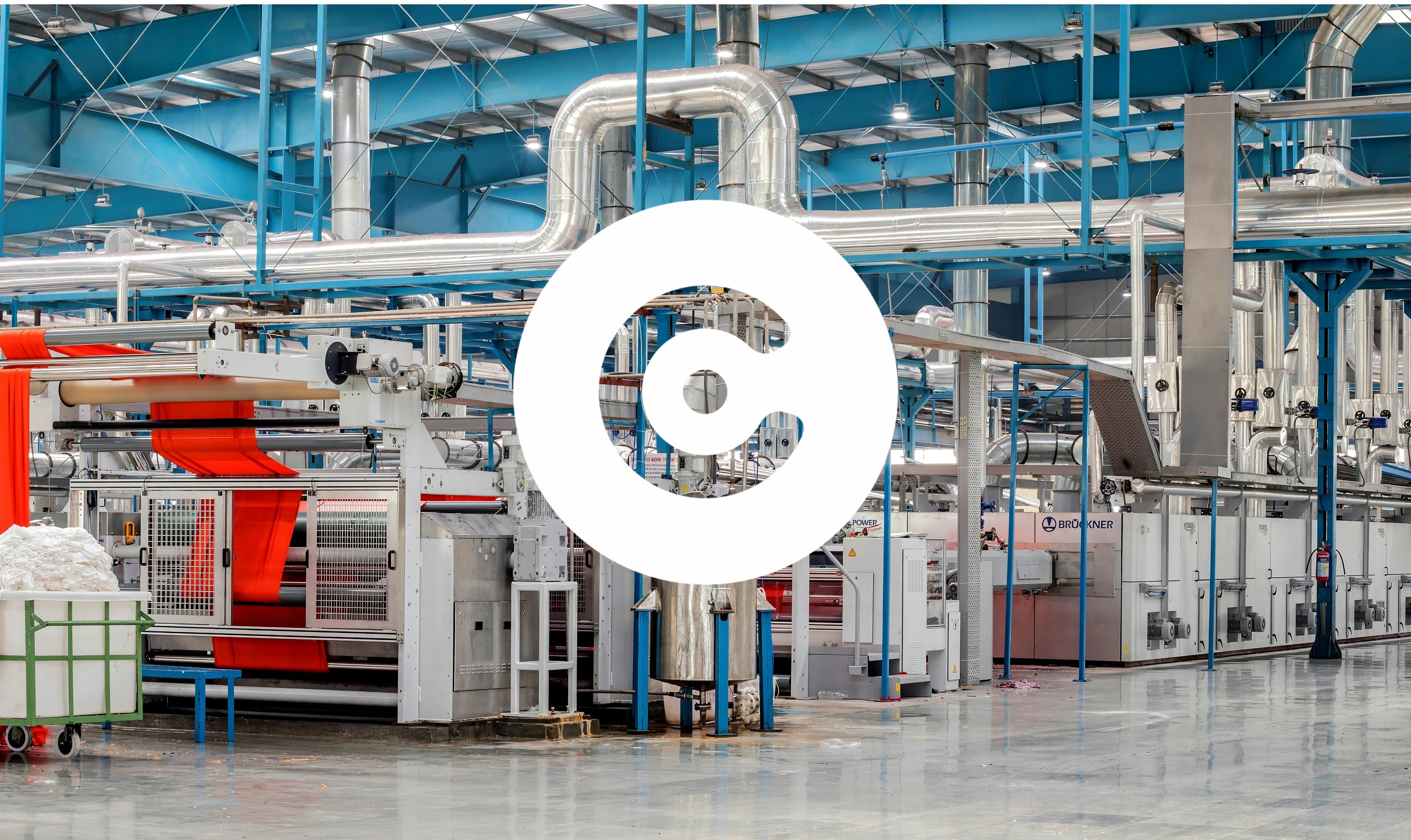 Industrial factory interior with large machinery, metal pipes, and a production line processing a wide red sheet of material.