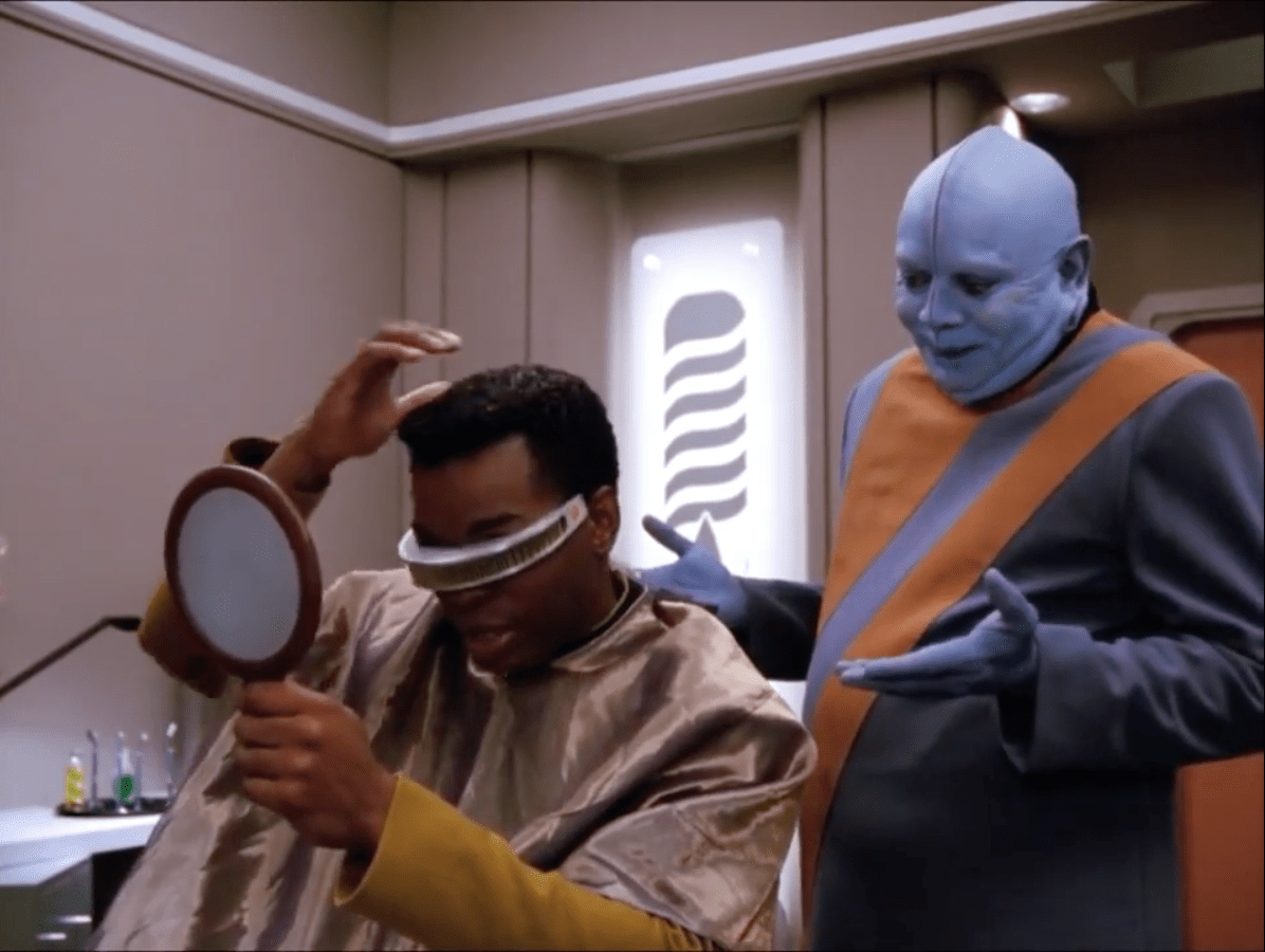 Screen capture from Star Trek: The Next Generation episode "Data's Day." In a futuristic barbershop aboard a starship, a seated black human male officer named Geordi Laforge, who is wearing a metallic visor that translates wavelengths of light into information his visual cortex can understand, examines his hair in a handheld mirror while sitting in a barber’s chair draped with a shiny silver cape. Standing beside him is his blue-skinned Bolian barber named Mott, dressed in a gray long sleeved tunic with a wide orange sash, gesturing animatedly as they discuss the hairstyle. The sleek, softly lit interior features curved walls and futuristic décor characteristic of the starship environment. Photo Credit: Streaming Service Paramount+.