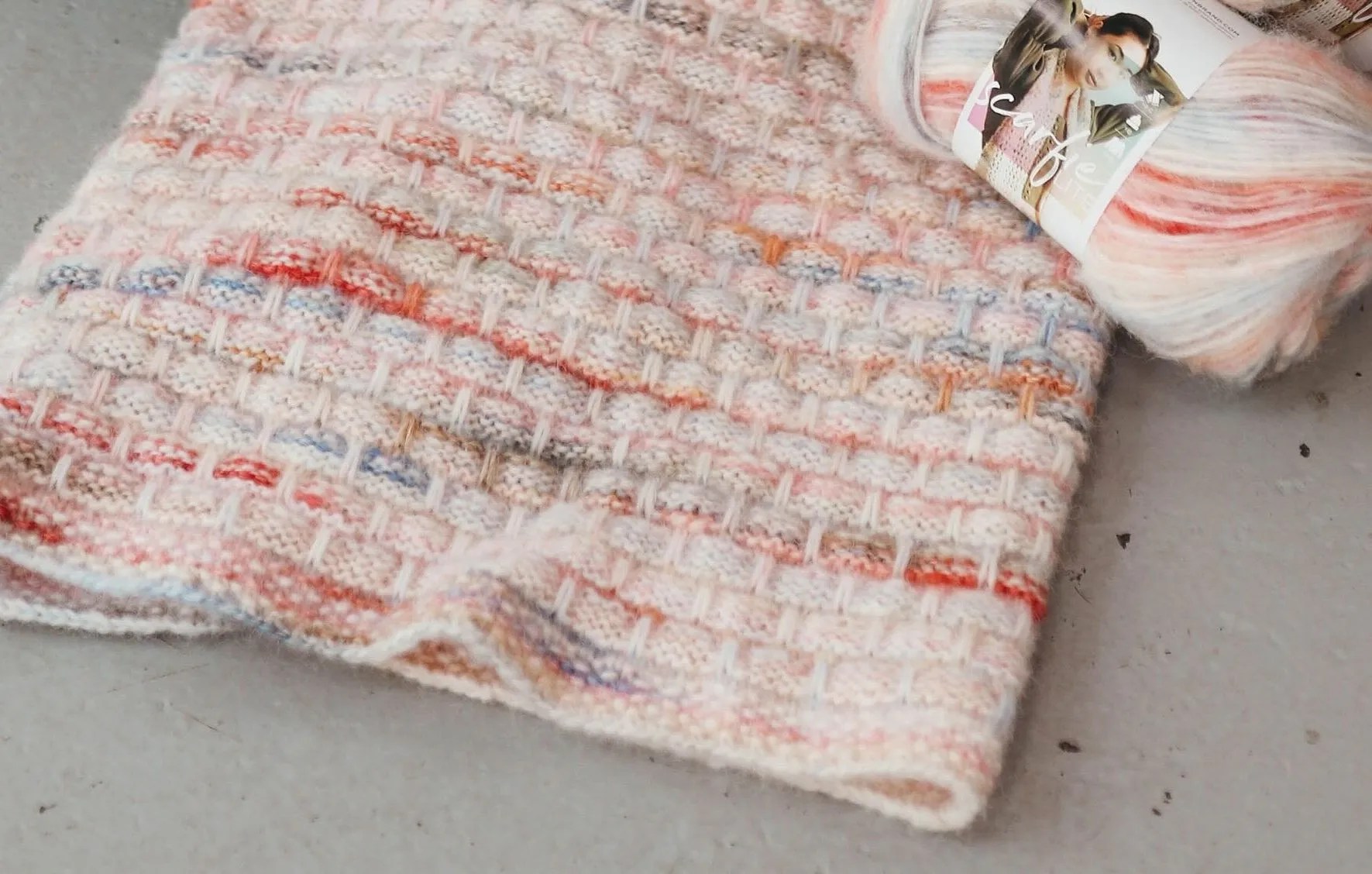 Blanket on the ground with a yarn skein of Scarfie lite laying next to it in the colorway Tartan