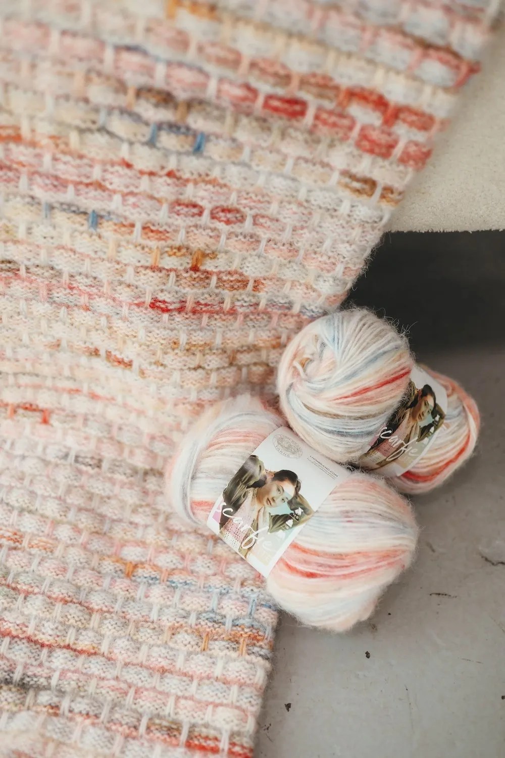Blanket laying on the ground with 2 skeins of yarn propped up against it. Blanket is red, blue, and cream.