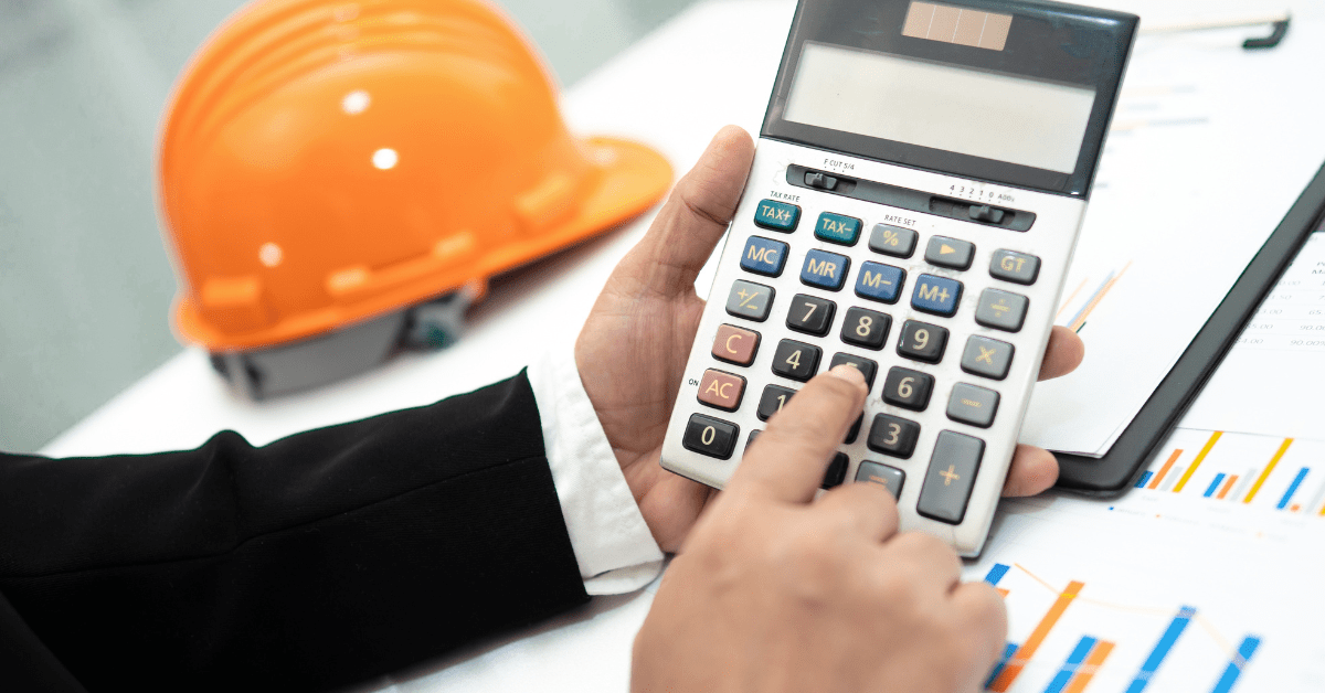 Construction accountant using a calculator with an orange hard hat and financial charts on the desk